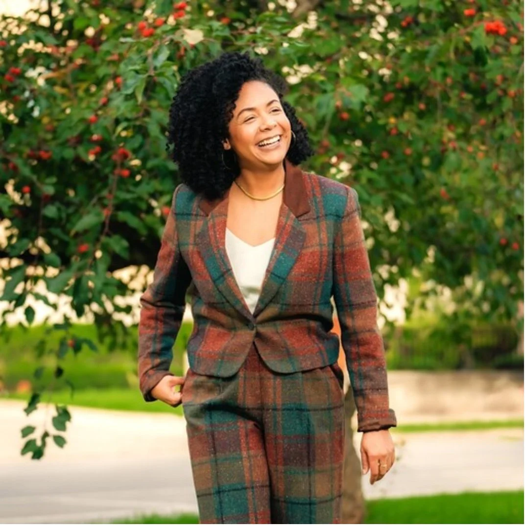 Smiling burnout recovery client, a woman, in plaid suit walking outdoors with trees in background.