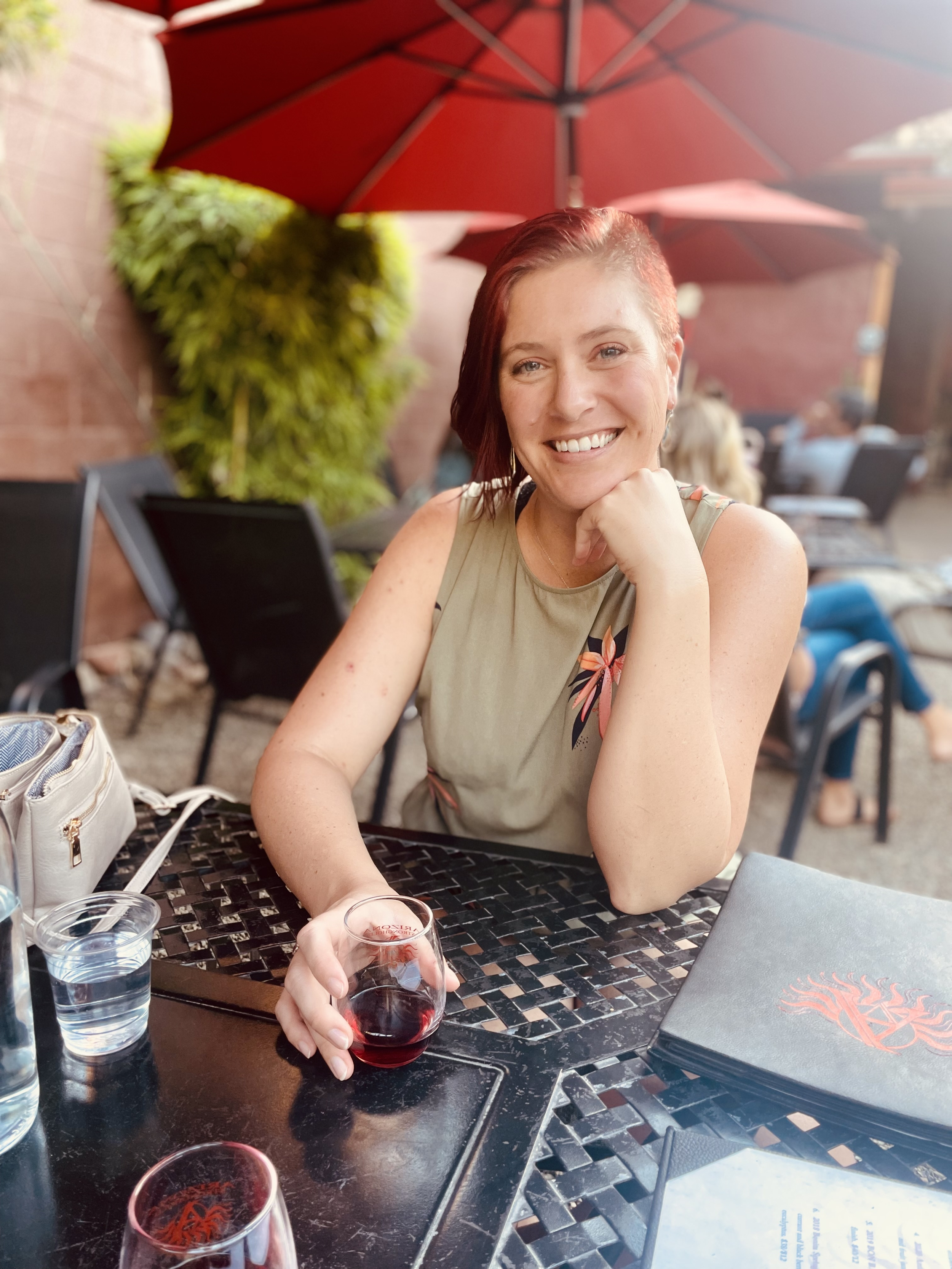 Burnout recovery client, a woman, seated at outdoor patio, holding a glass of red wine, with red umbrellas and greenery in the background.