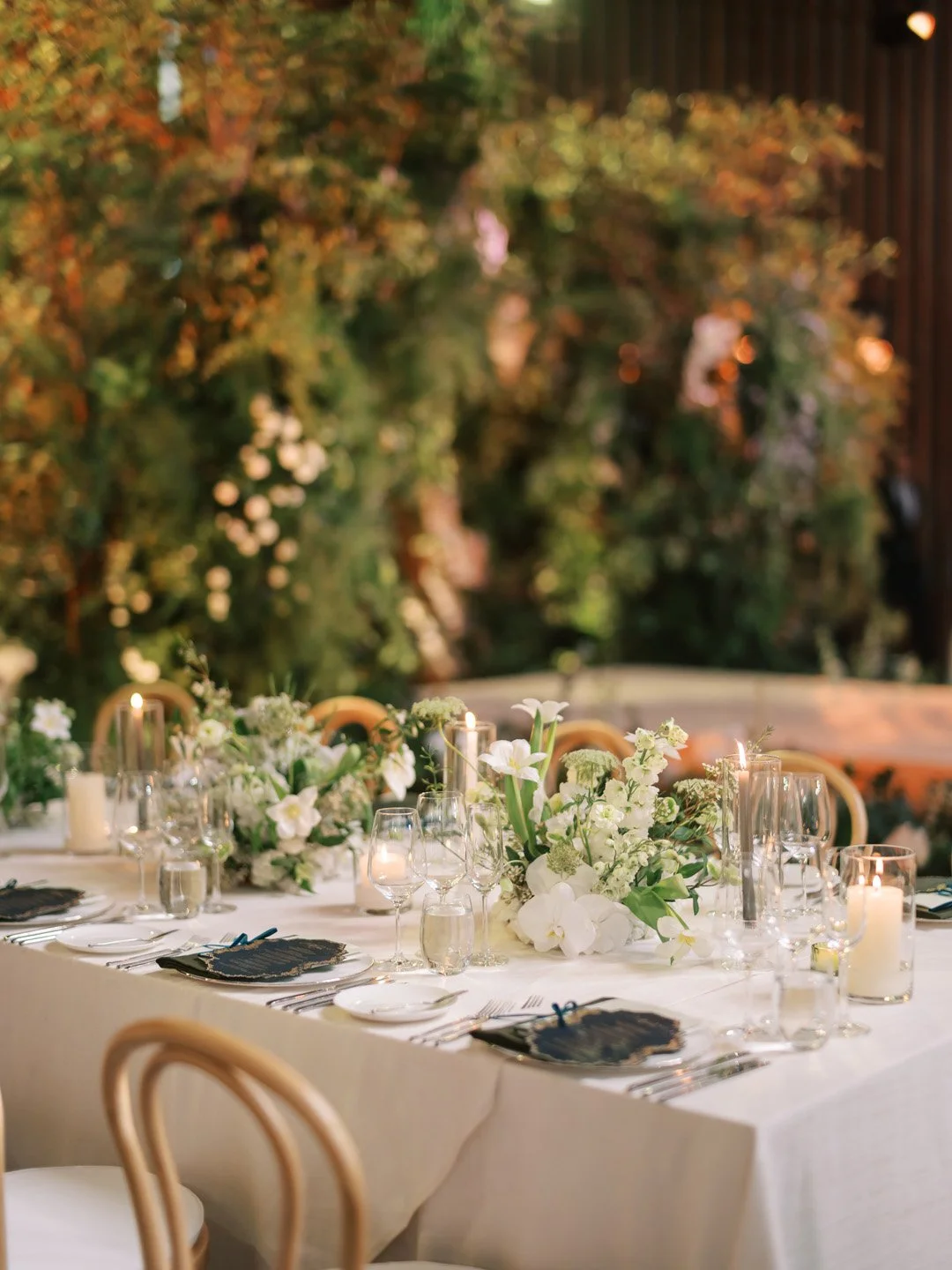 Elegant dining table with white floral centerpiece, candles, wine glasses, and place settings in a warmly lit, outdoor garden setting with blurred greenery in the background.