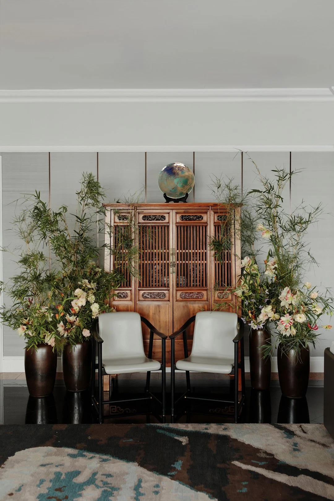 A living room corner with two white chairs facing each other, flanked by large dark vases filled with green foliage and white flowers. Behind, a wooden cabinet with carved details and a globe on top is visible. The wall is painted light gray.