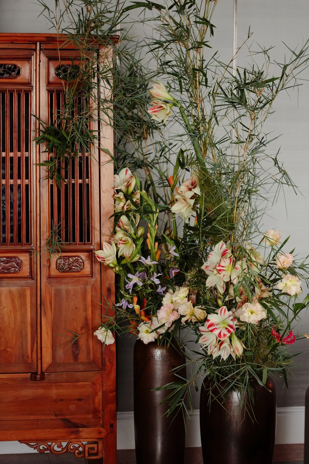 Two tall brown vases filled with an arrangement of light-colored flowers and greenery, placed next to a wooden cabinet with intricate carvings.