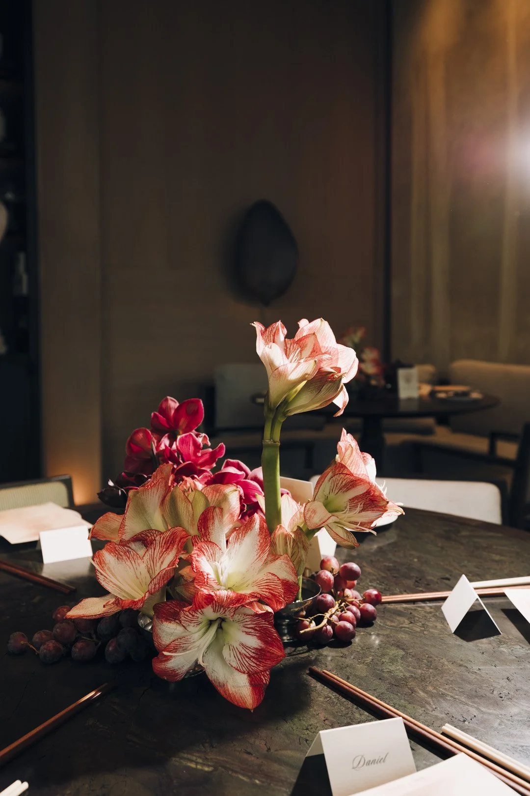 A floral centerpiece with large pink and white amaryllis flowers and red grapes on a dark dining table in a dimly lit restaurant.