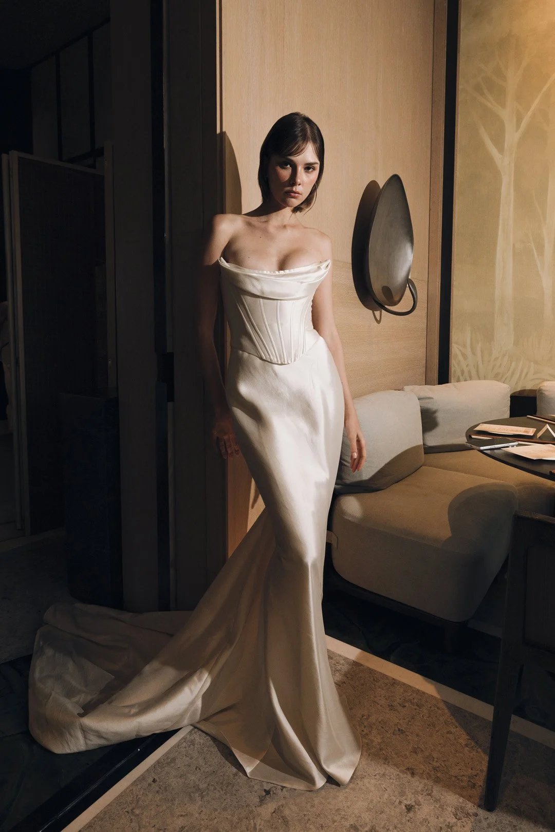 A woman in a long, off-shoulder, satin dress standing in a dimly lit room with wooden walls, a sitting area, and decorative wall art.