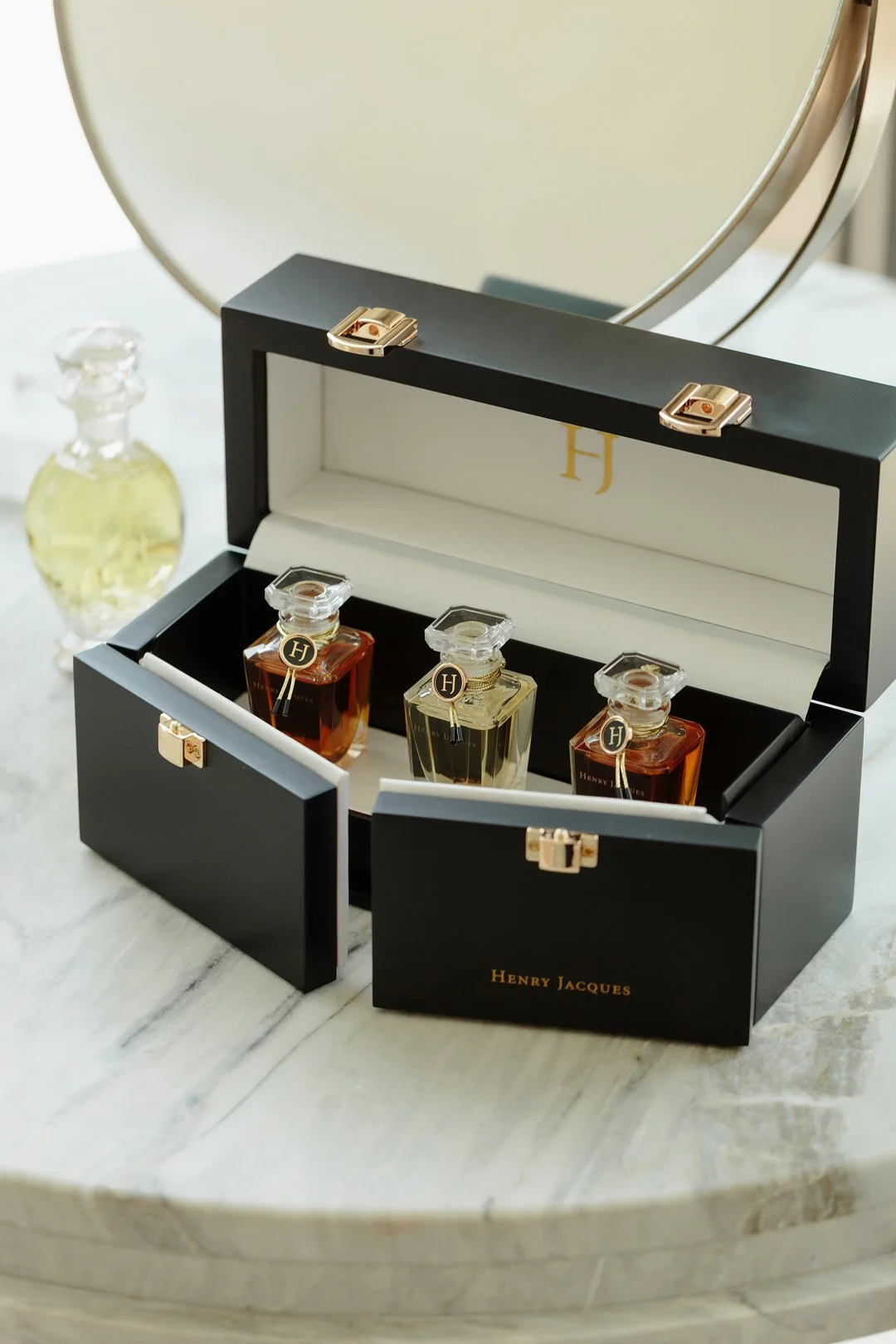 A black and white Henry Jacques perfume box containing three small glass perfume bottles with gold and black tags, placed on a marble table. A round mirror and a glass perfume bottle with yellow perfume are in the background.