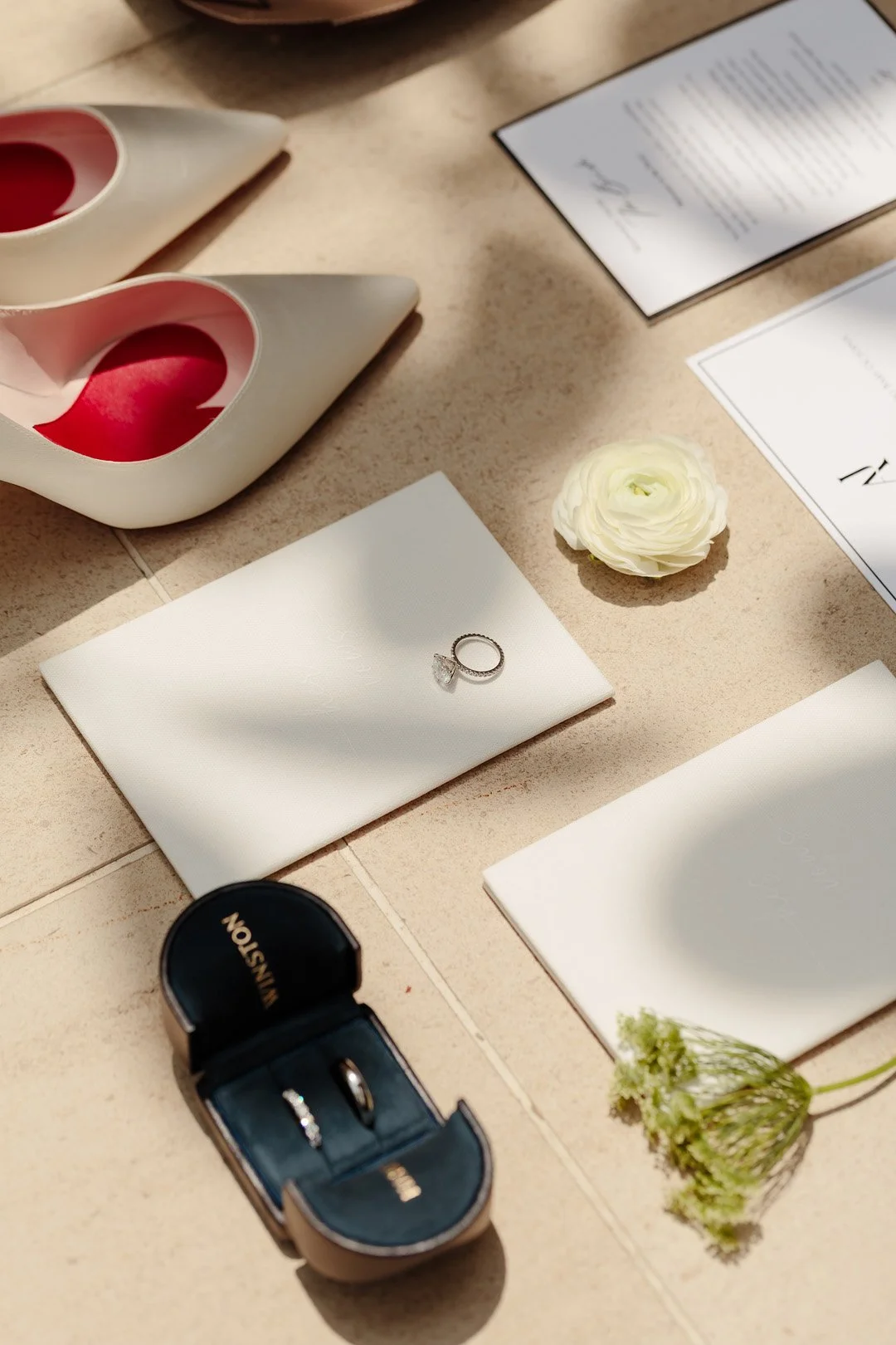 Wedding ring on a white napkin with a blue jewelry box, surrounded by white cards, a white flower, and decorative objects on a beige surface.