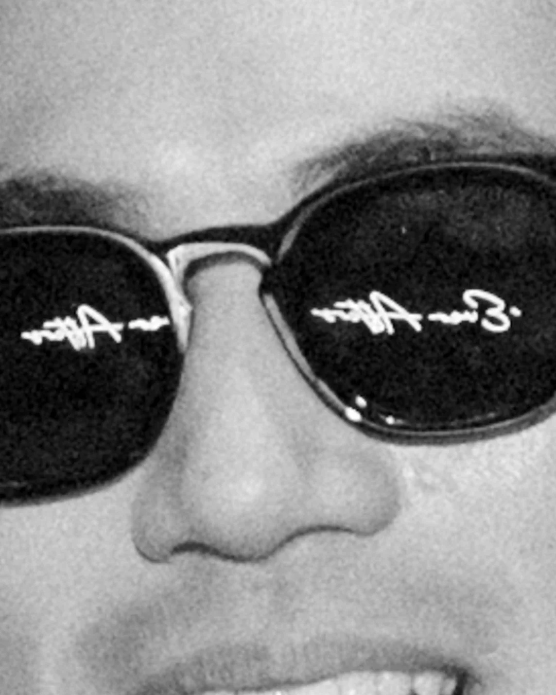Close-up of a person's face wearing round sunglasses, with partially visible nose and mouth, in black and white.