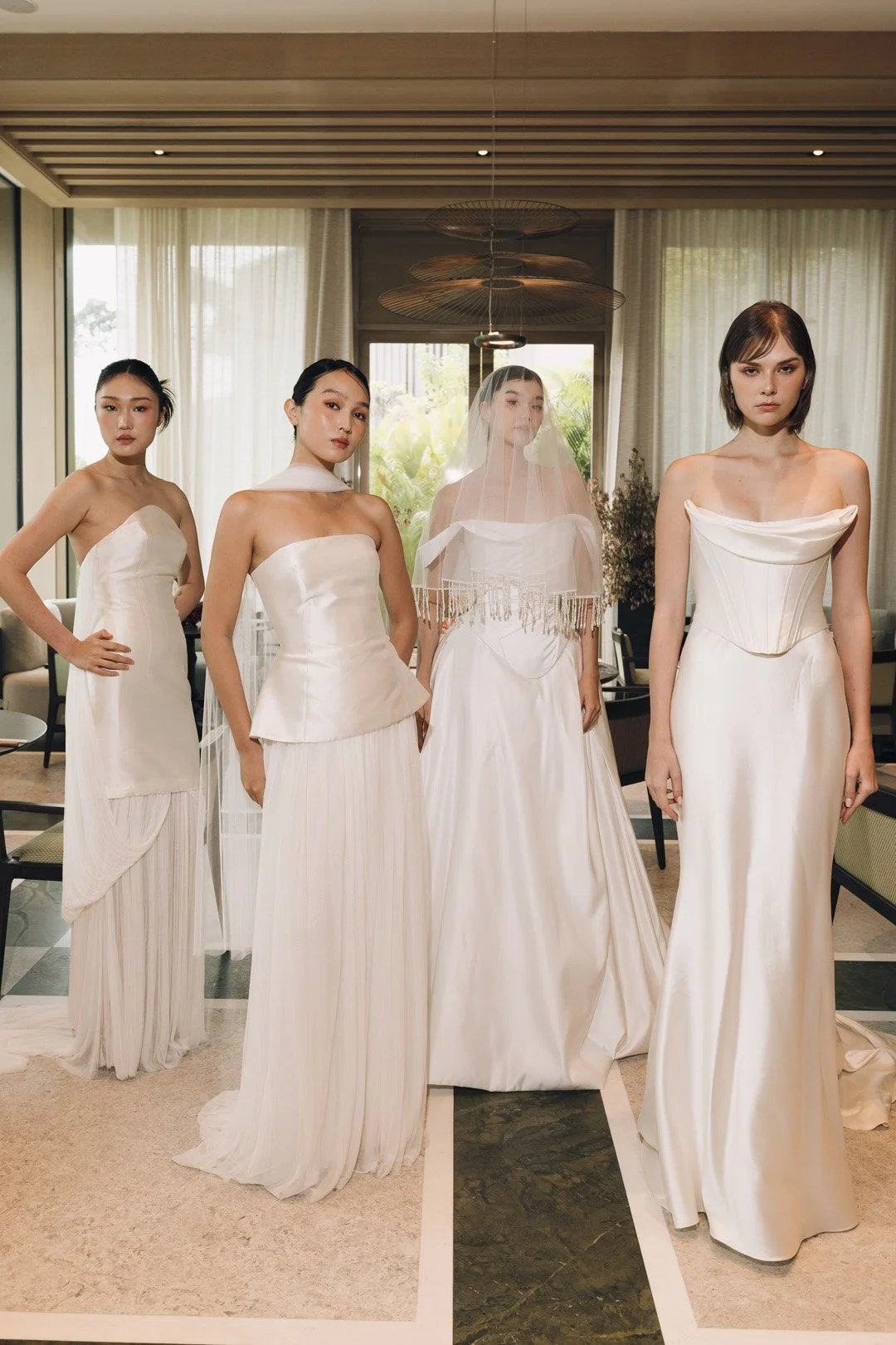 Five women standing in a room with large windows, all wearing elegant white dresses, with one woman in the center wearing a veil, suggesting a bridal or fashion photoshoot.