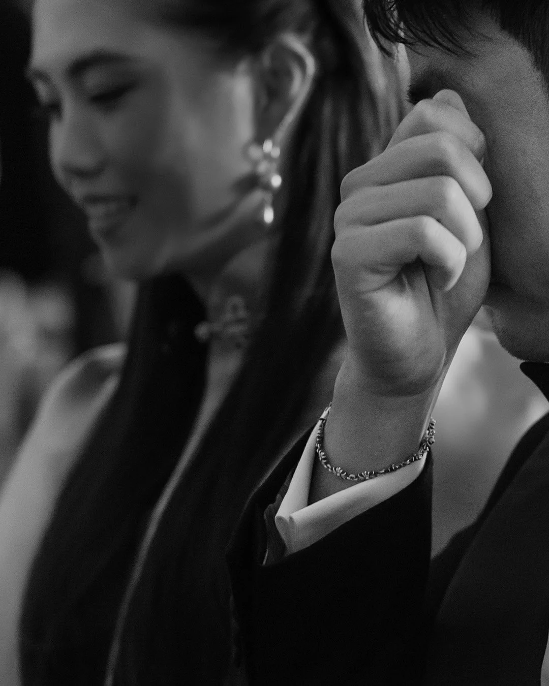 A black and white photo of two people sitting closely; the woman is smiling and looking down, wearing earrings, and the man is resting his head on his hand with a bracelet on his wrist.