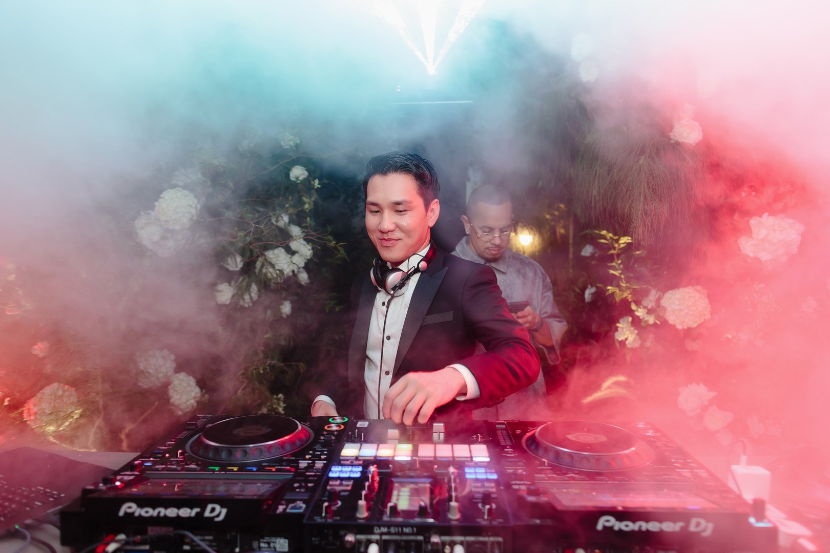 A young male DJ in a tuxedo with headphones around his neck is DJing at an event, with a background of flowers and ambient lighting, and a person in the background using a smartphone.