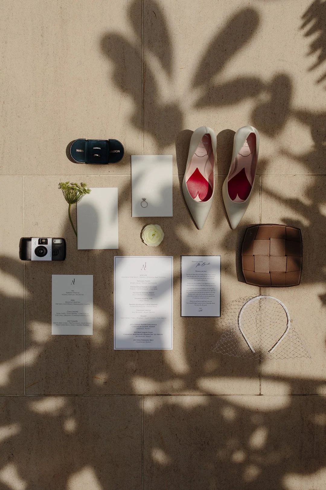 Flat lay of wedding items on a tiled floor, including white high-heeled shoes with pink insoles, a small brown basket, a white headband with mesh veil, a decorative flower, a ring box with a ring, a camera, a vintage film camera, handwritten wedding 
