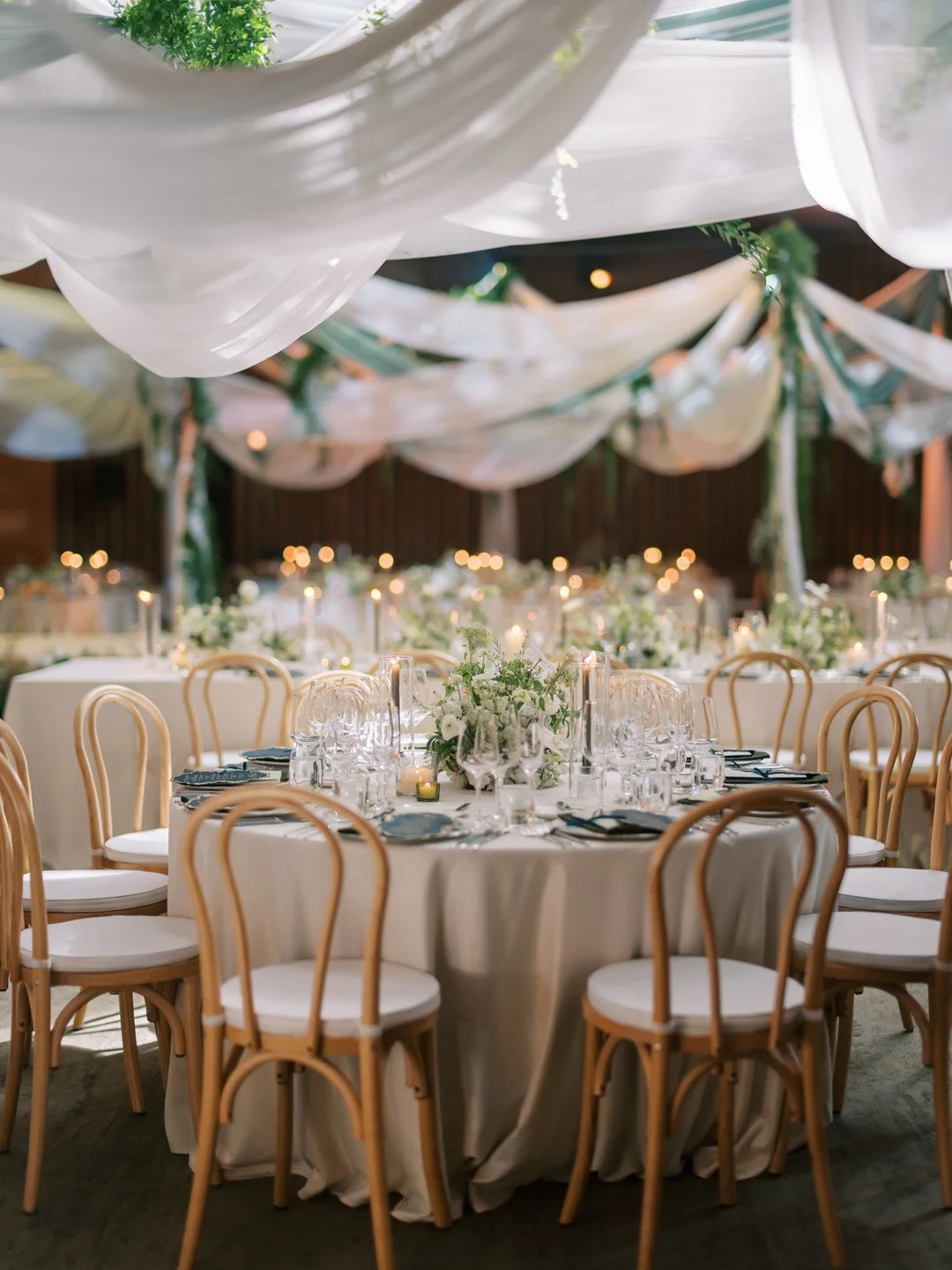 Elegant event dining table with floral centerpieces, surrounded by wooden chairs with white cushions, decorated with draped fabric and soft lighting.