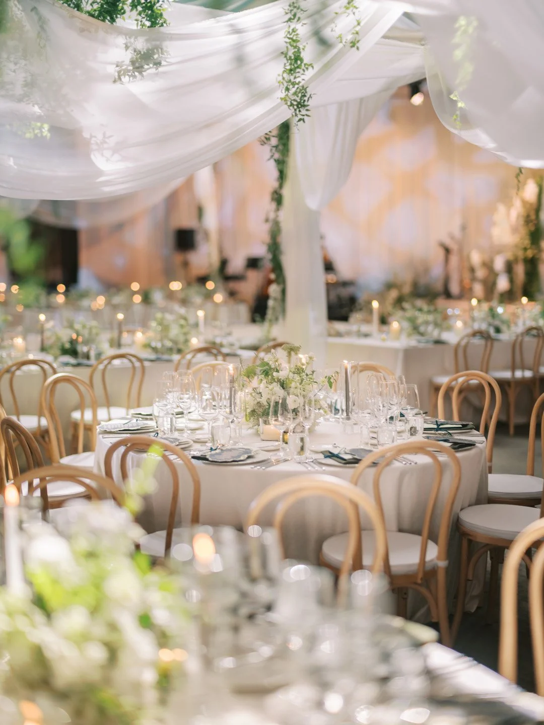 Elegant banquet setup with round tables, white tablecloths, floral centerpieces, and wooden chairs in a decorated indoor venue with draped fabric and soft lighting.