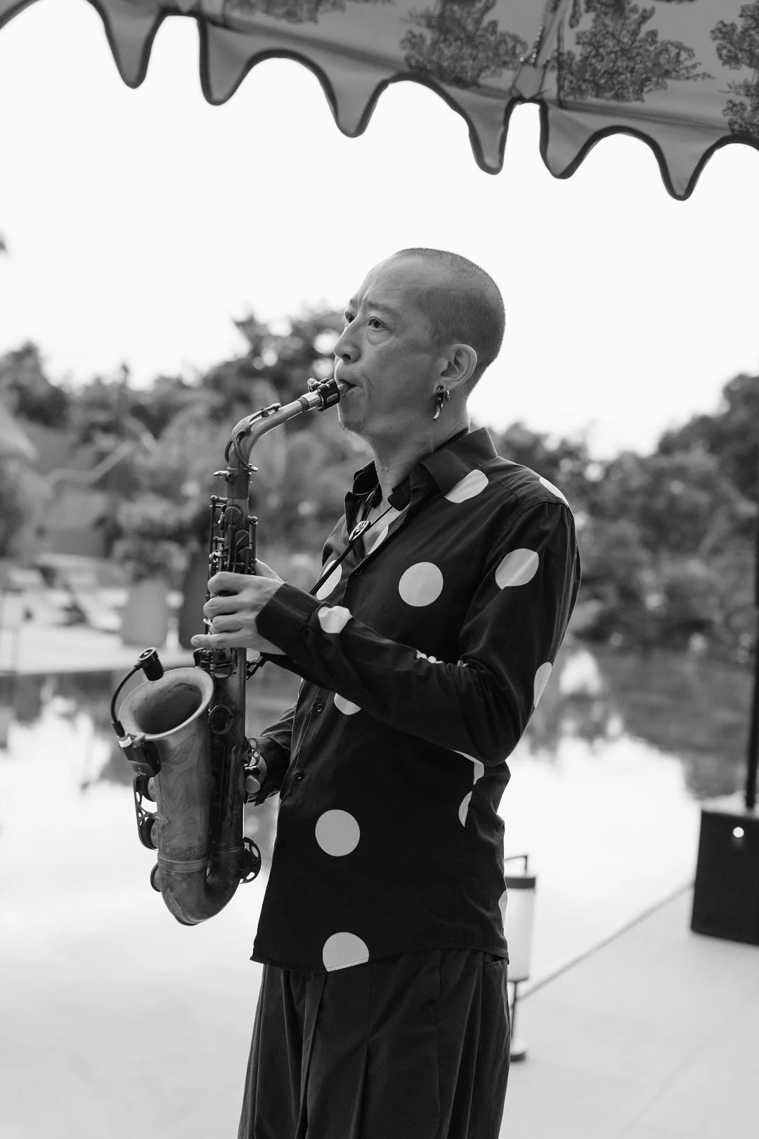 A person playing the saxophone outdoors under a canopy, wearing a polka dot shirt and earrings, with trees and water in the background.
