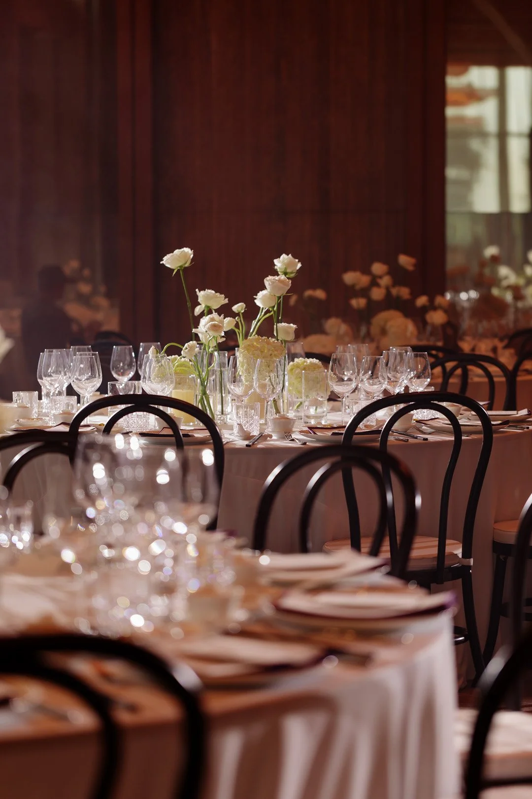 Elegant banquet table with white floral centerpieces, surrounded by black chairs, set with glassware and plates, in a warmly lit event hall.