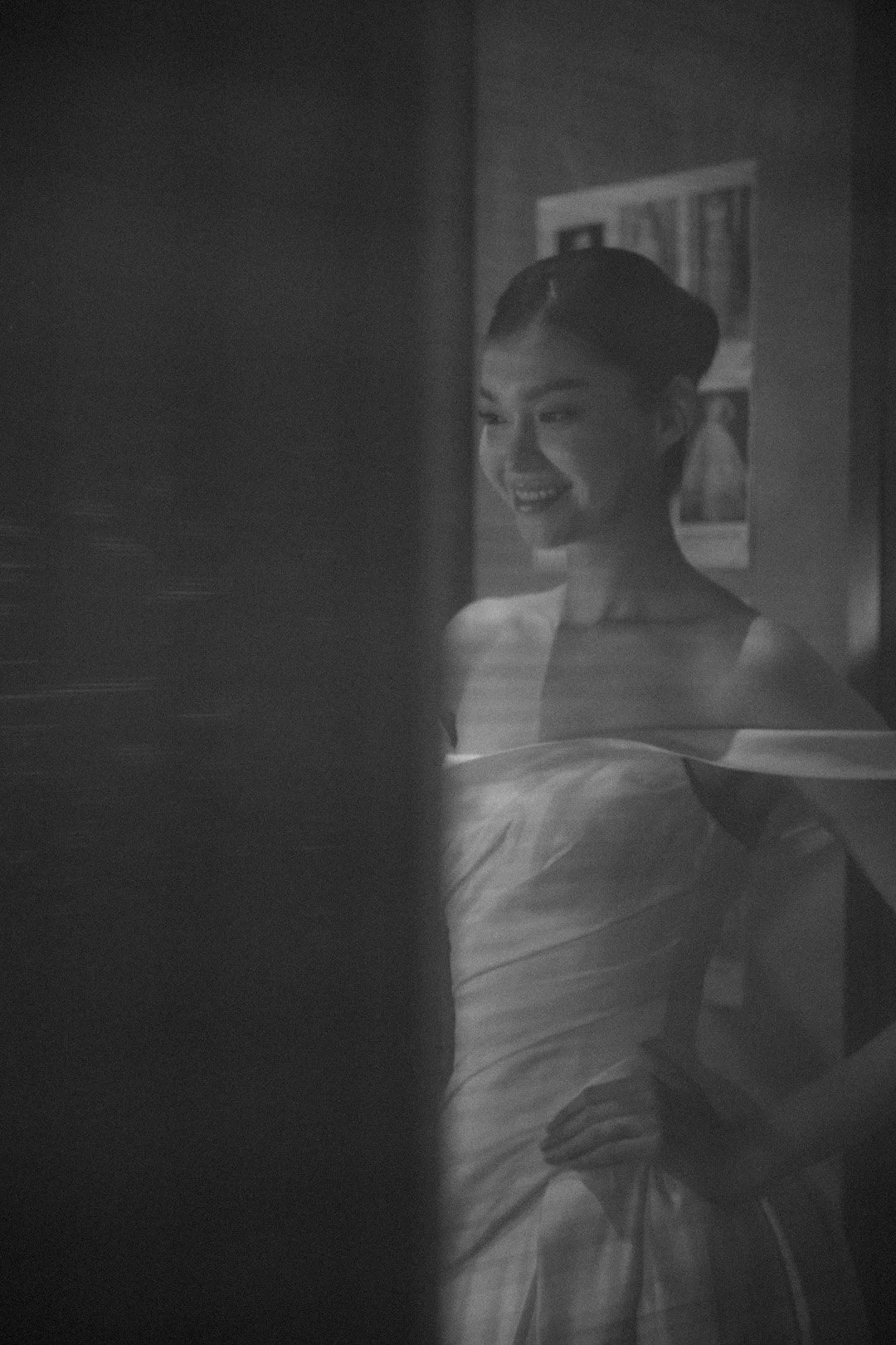 A black and white photo of a young woman with a traditional hairstyle, smiling and looking to her right, partially obscured by a vertical object in the foreground.