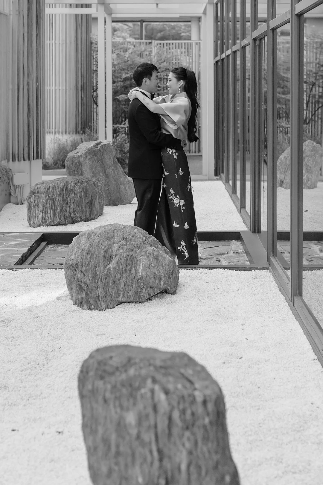 A couple in formal attire embracing inside a modern glass-walled space with rocks and white gravel on the ground.
