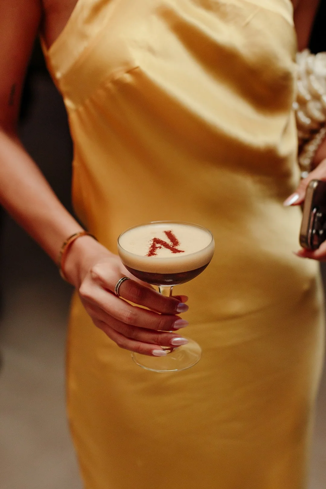 Person holding a cocktail glass with a dark drink topped with foam, garnished with a red letter 'N' drawn in cinnamon or cocoa powder.