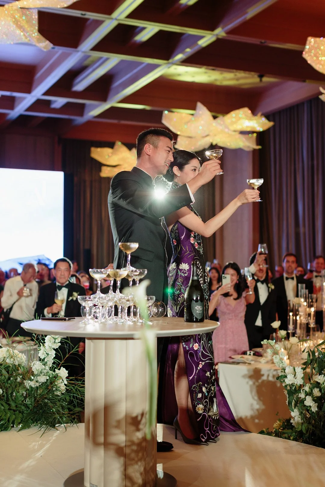 A bride and groom celebrating at their wedding reception, raising champagne glasses in a toast, with guests watching and taking photos in the background.