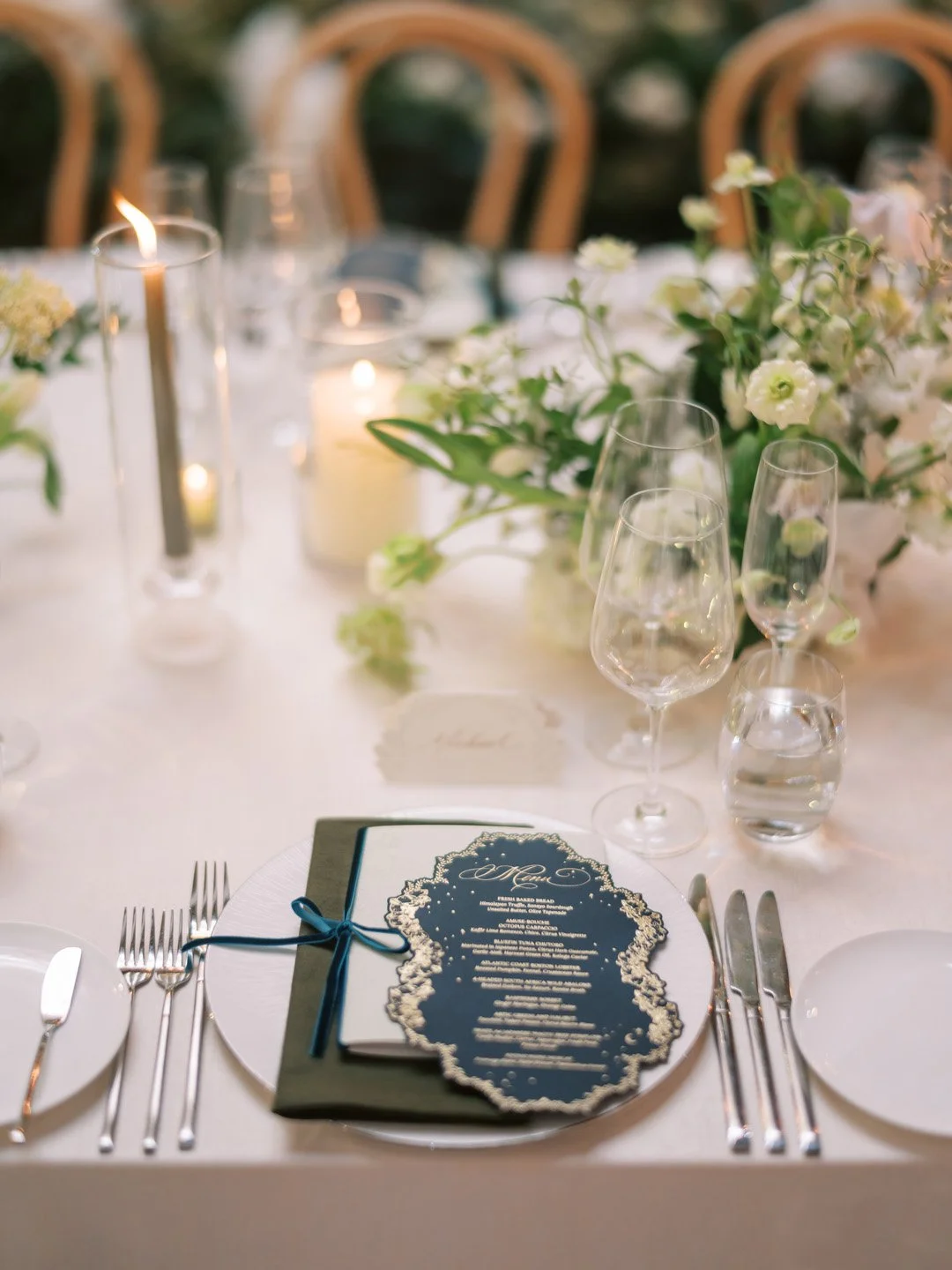 Elegant table setting with a dark blue menu, wine glasses, water glasses, floral centerpiece with white flowers, and lit candles.