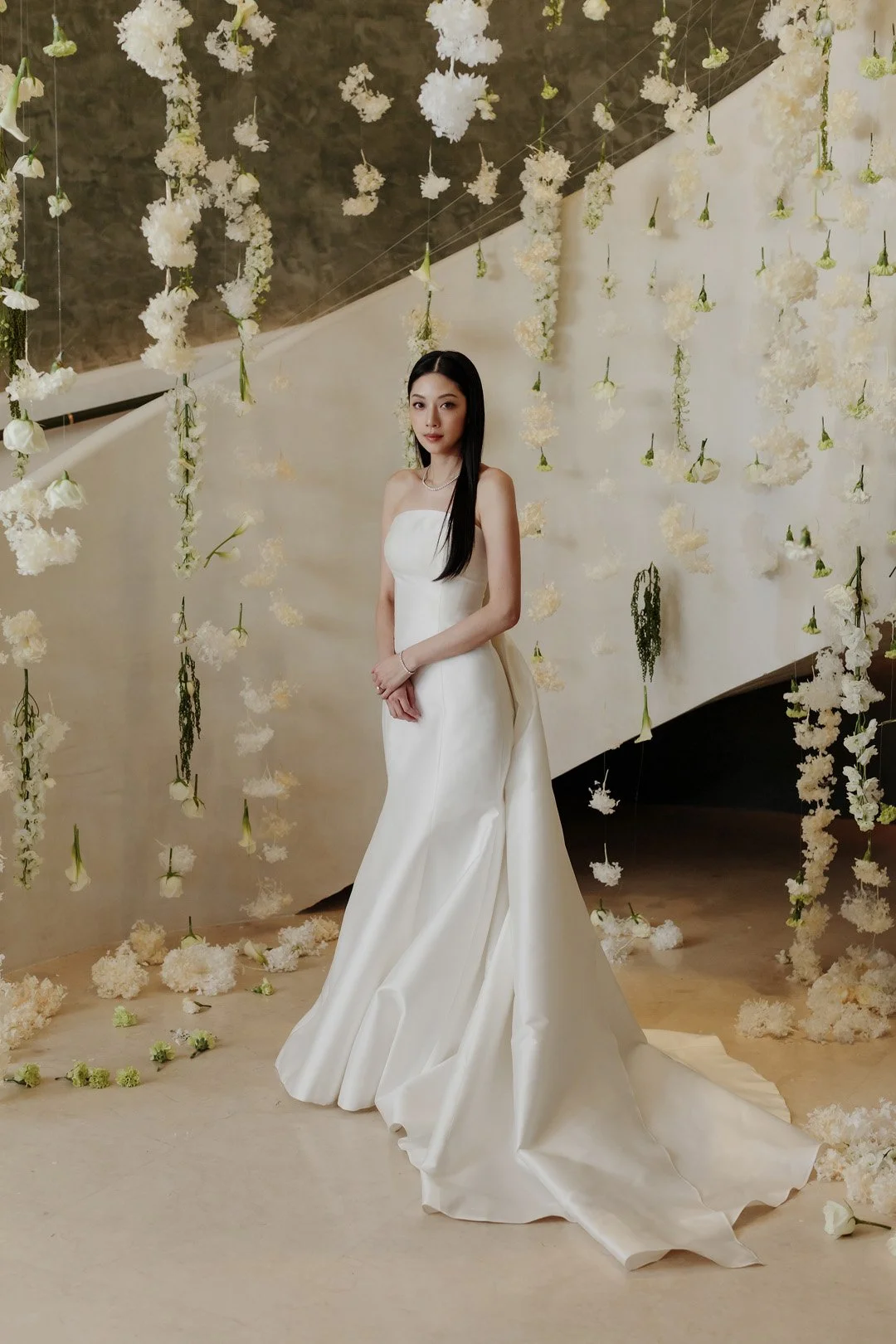 A woman in a strapless white wedding dress standing in front of hanging white flowers and a decorated backdrop.