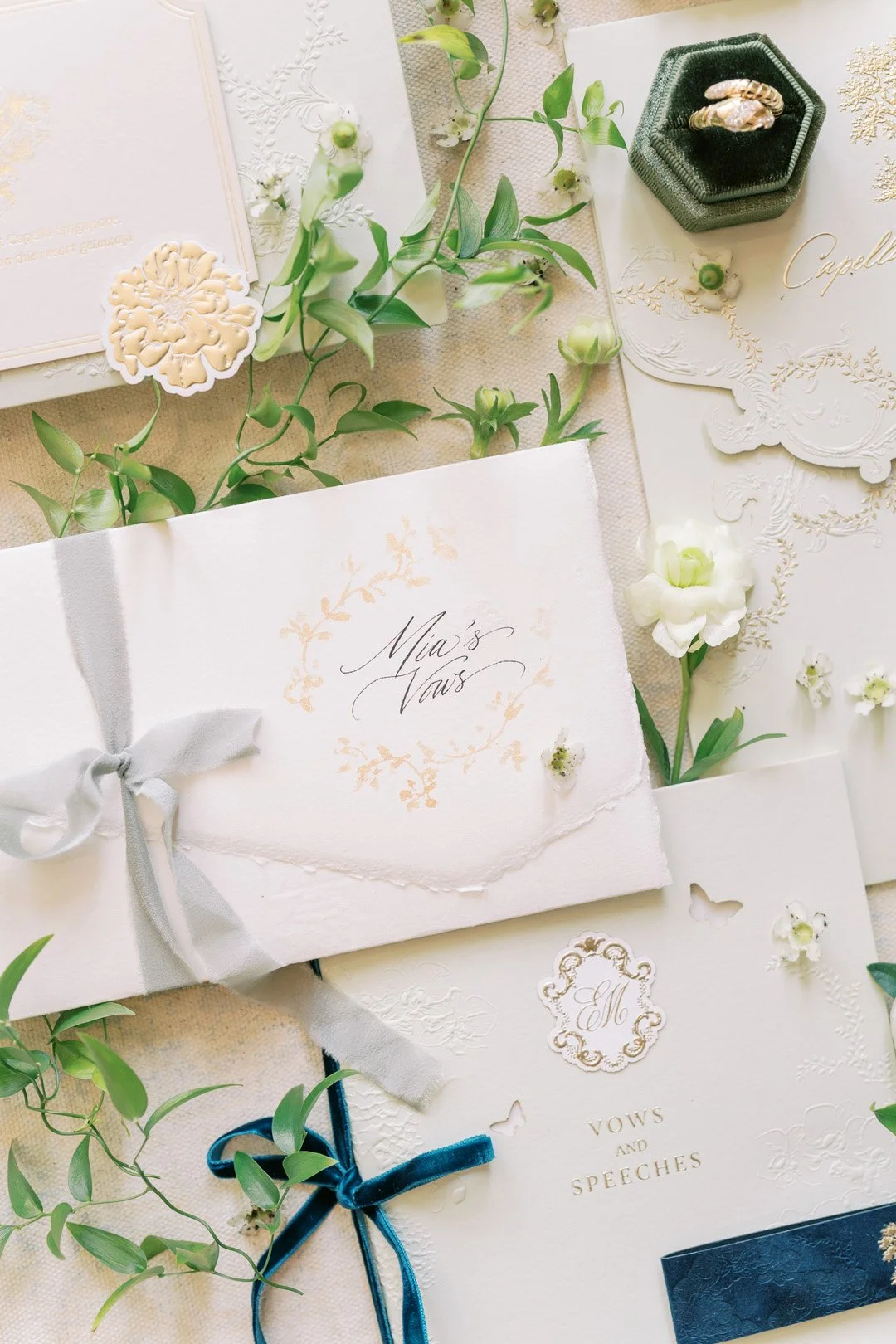 Wedding invitation suite with floral embellishments, featuring an envelope labeled "Mia's Vows" and another labeled "Vows and Speeches," surrounded by white flowers, greenery, and decorative accents.