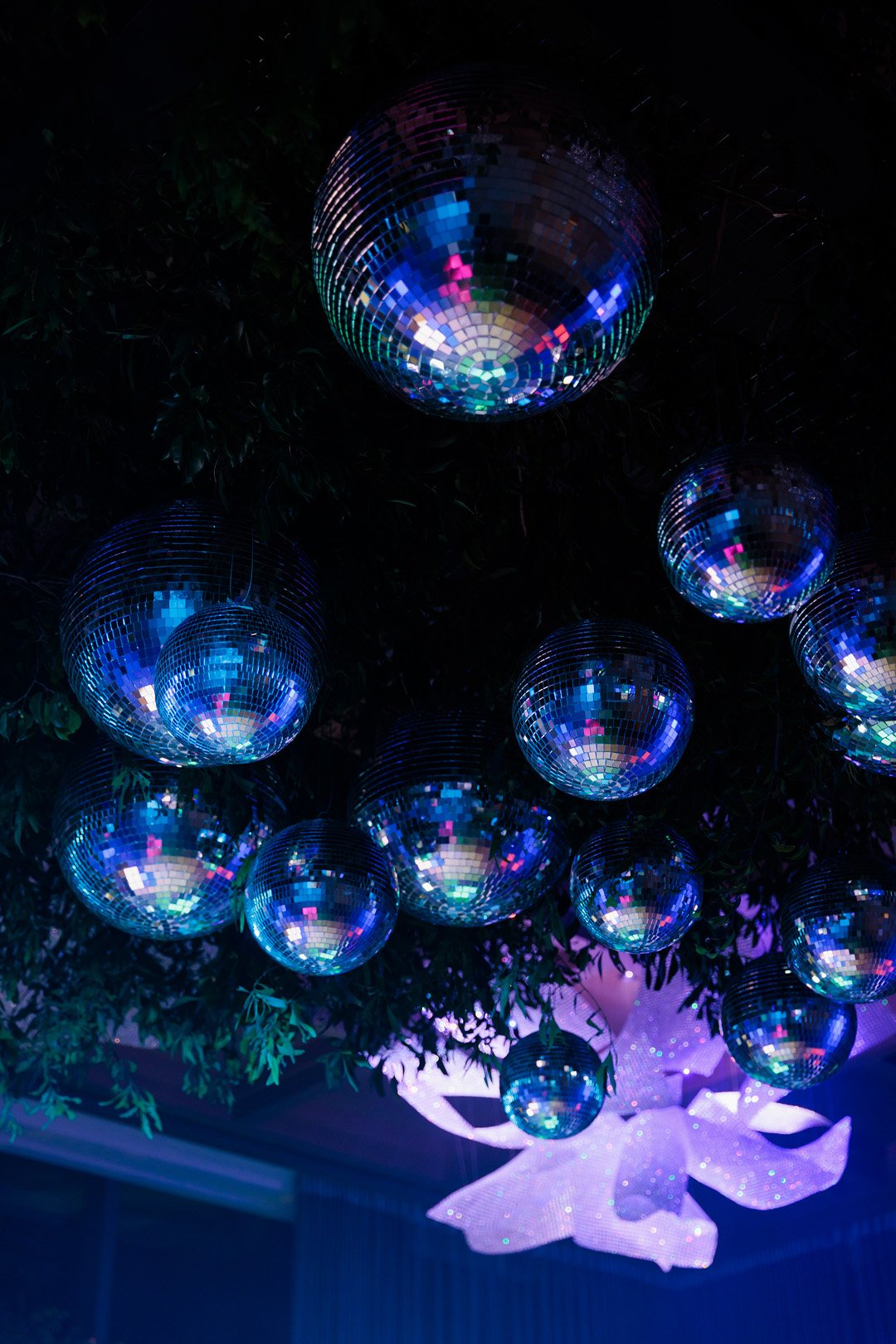 Multiple disco balls of various sizes hanging from a ceiling against a dark background, reflecting colorful lights, with a large white bow decoration underneath.