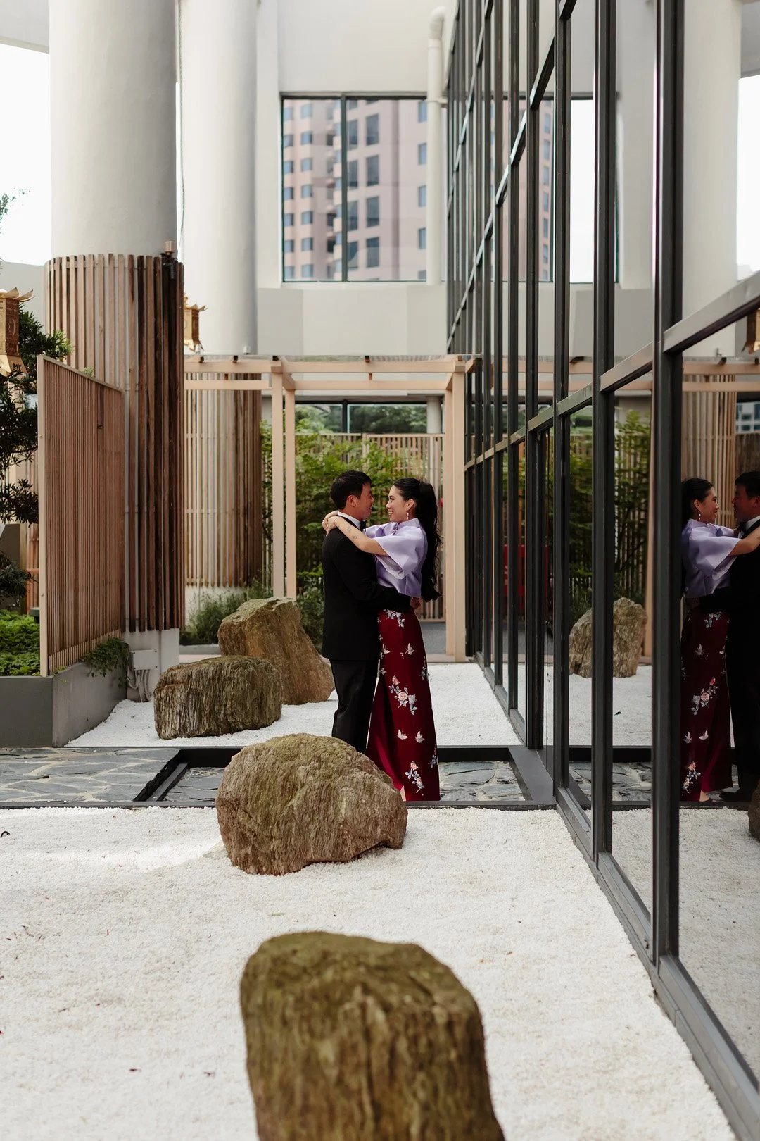 A couple dressed in formal attire dancing and smiling in a modern indoor space with large glass windows, rocks, and minimalist decor.
