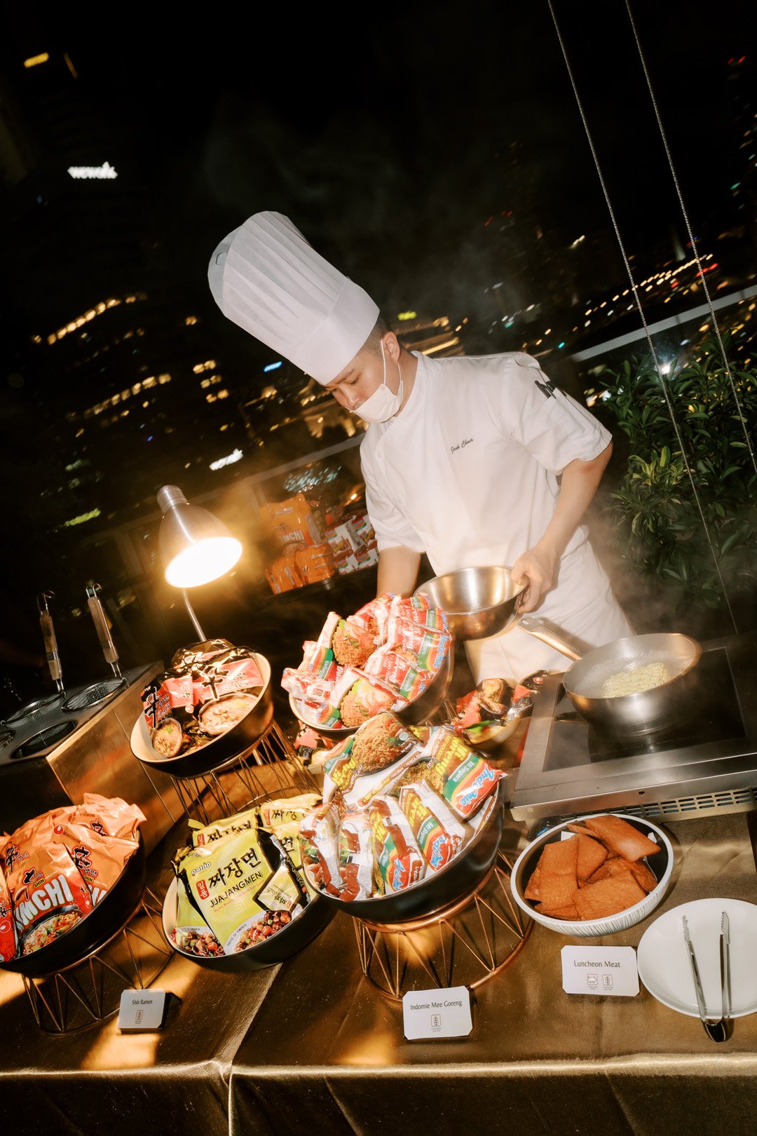 A chef wearing a white chef's hat, mask, and uniform is preparing food on a self-serve station with instant noodles, packaged snacks, and luncheon meat. The station is lit with a small desk lamp, and city lights are visible through the window in the 