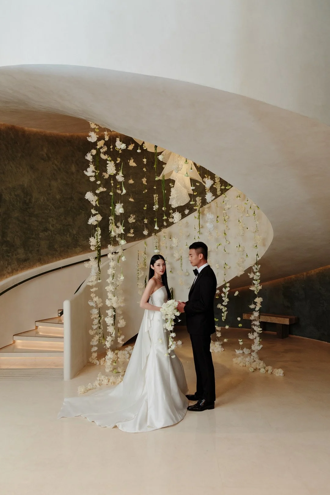 Young couple in wedding attire standing in a modern, indoor space decorated with white flowers and hanging floral arrangements. The bride wears a white wedding gown and holds a bouquet, and the groom is in a black tuxedo. They face each other, holdin