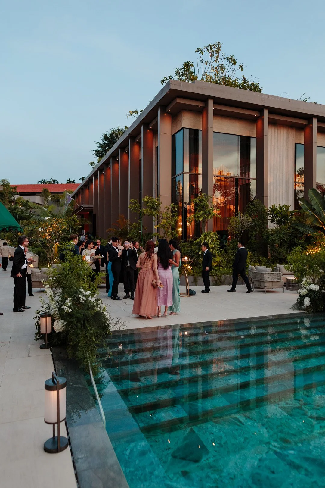 An outdoor evening event with people dressed in formal attire gathering near a modern building with glass windows, surrounded by lush greenery, next to a swimming pool.
