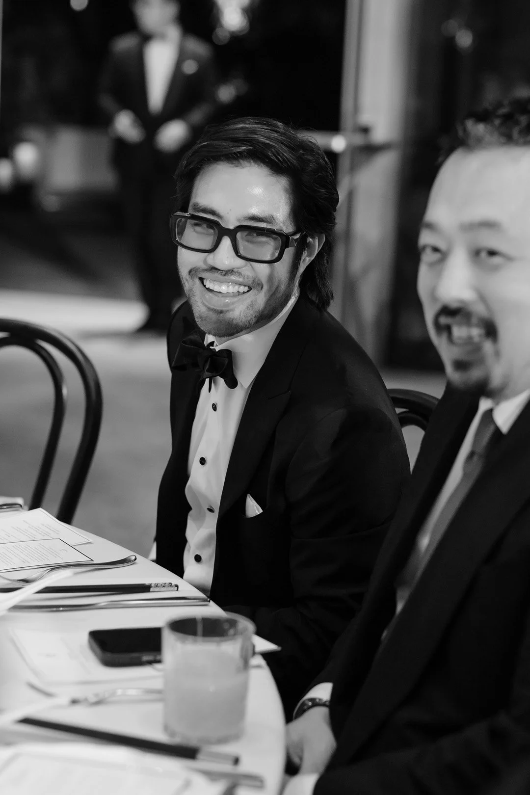 A smiling man wearing glasses, a tuxedo, and a bow tie, sitting at a formal dinner table with another man in a tuxedo, in a black-and-white photo.