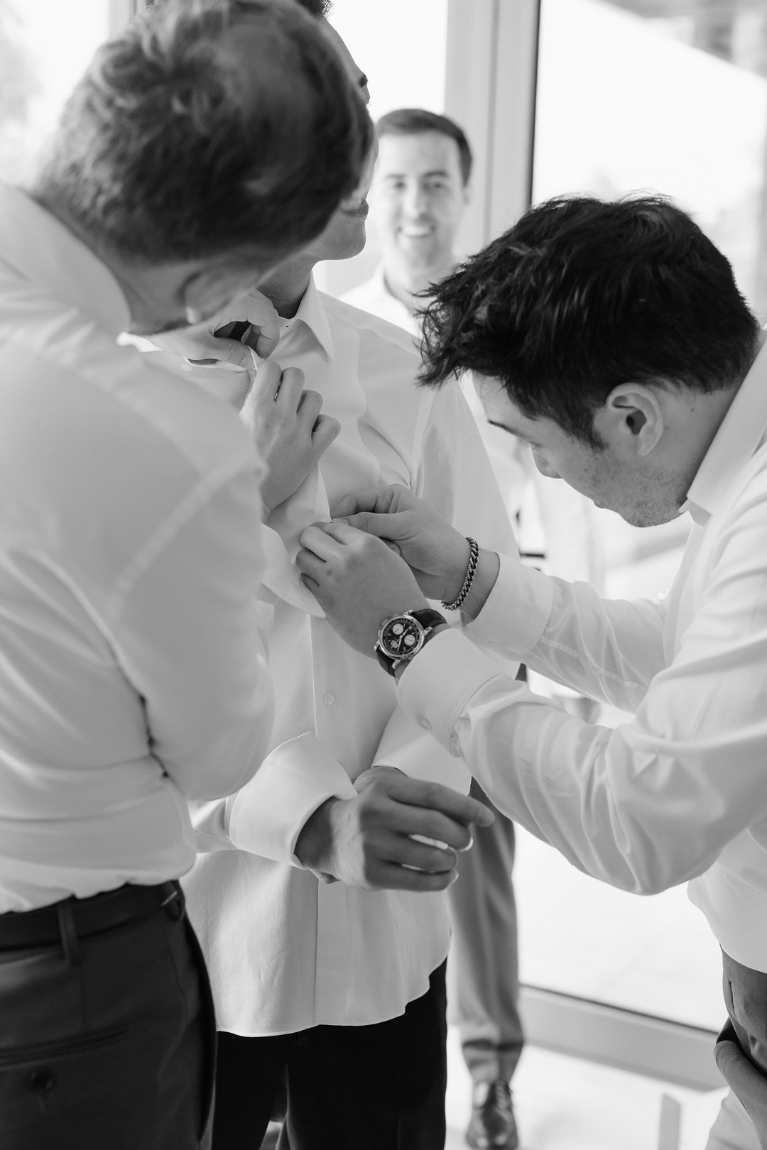 Young man assisting in cuffing another man's shirt sleeve during a formal occasion with other men in the background.