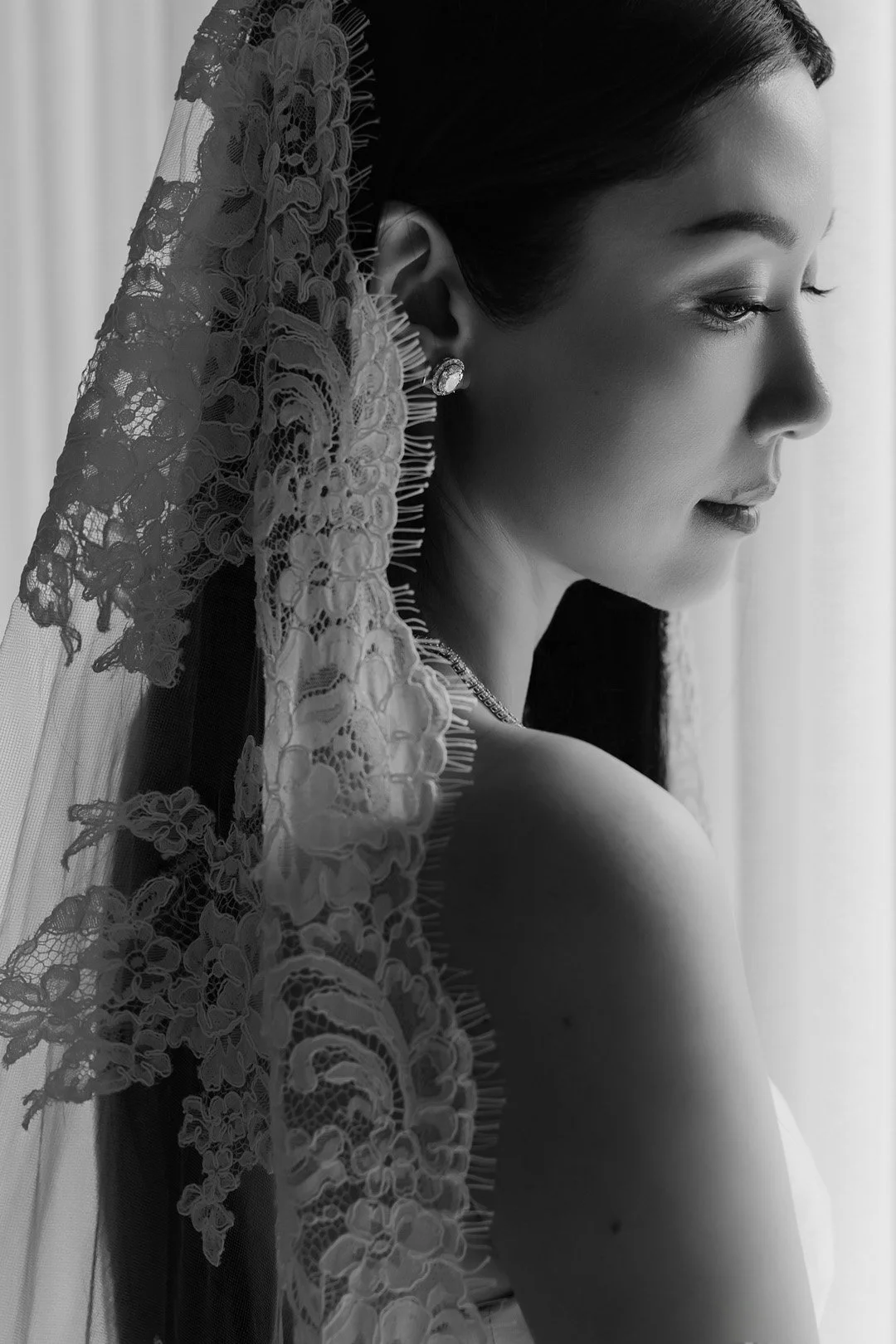 Black and white photo of a woman with a lace veil, wearing earrings and a necklace, looking out a window with a serene expression.