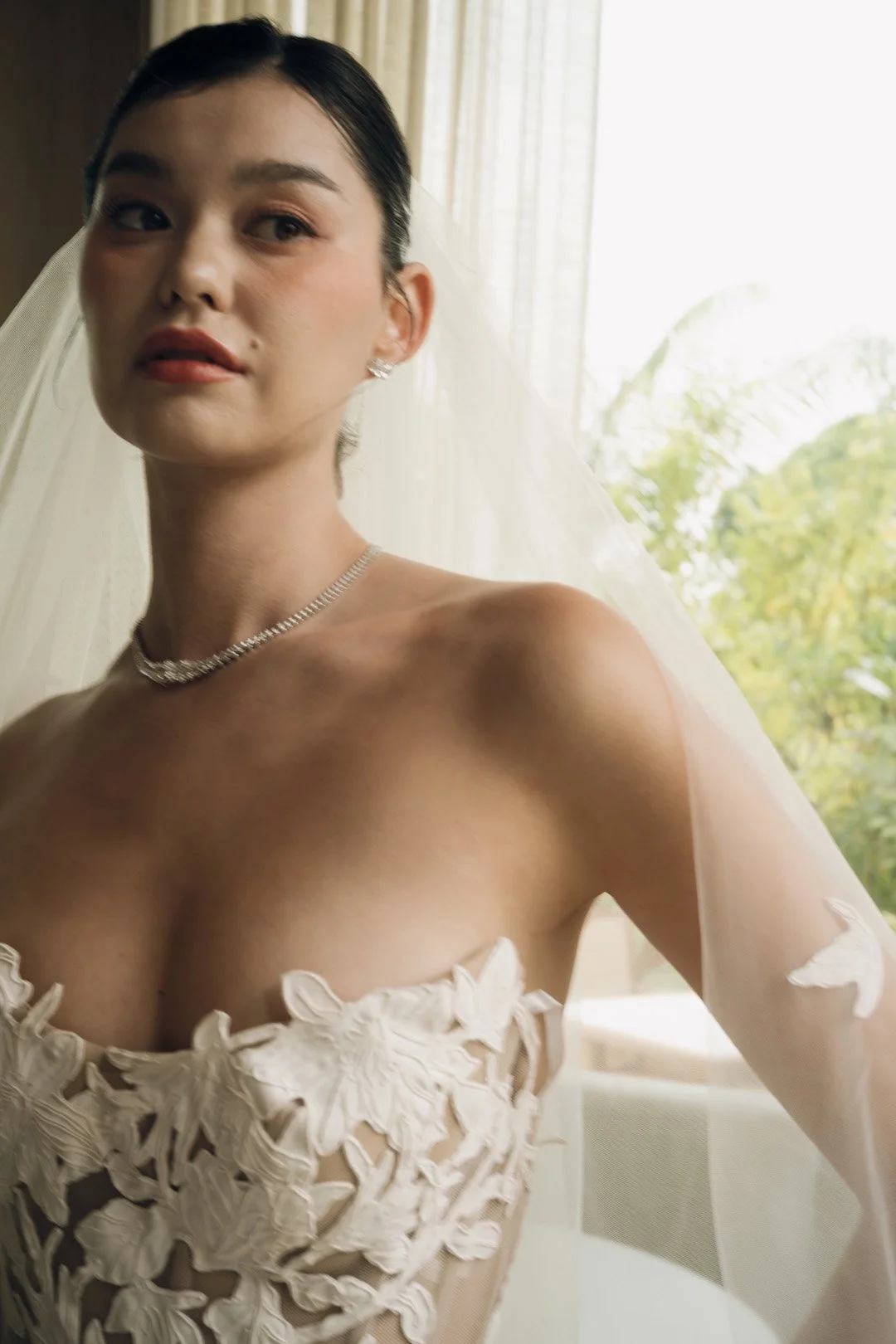 Close-up of a woman in a wedding dress with floral embroidery, veil, and jewelry, standing near a window with greenery outside.