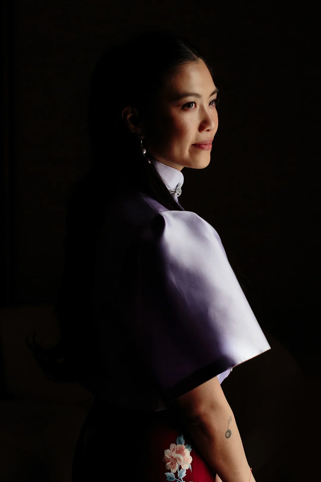 A woman with dark hair in a braid, wearing a purple silk top with a high collar, looking to the side, dark background.