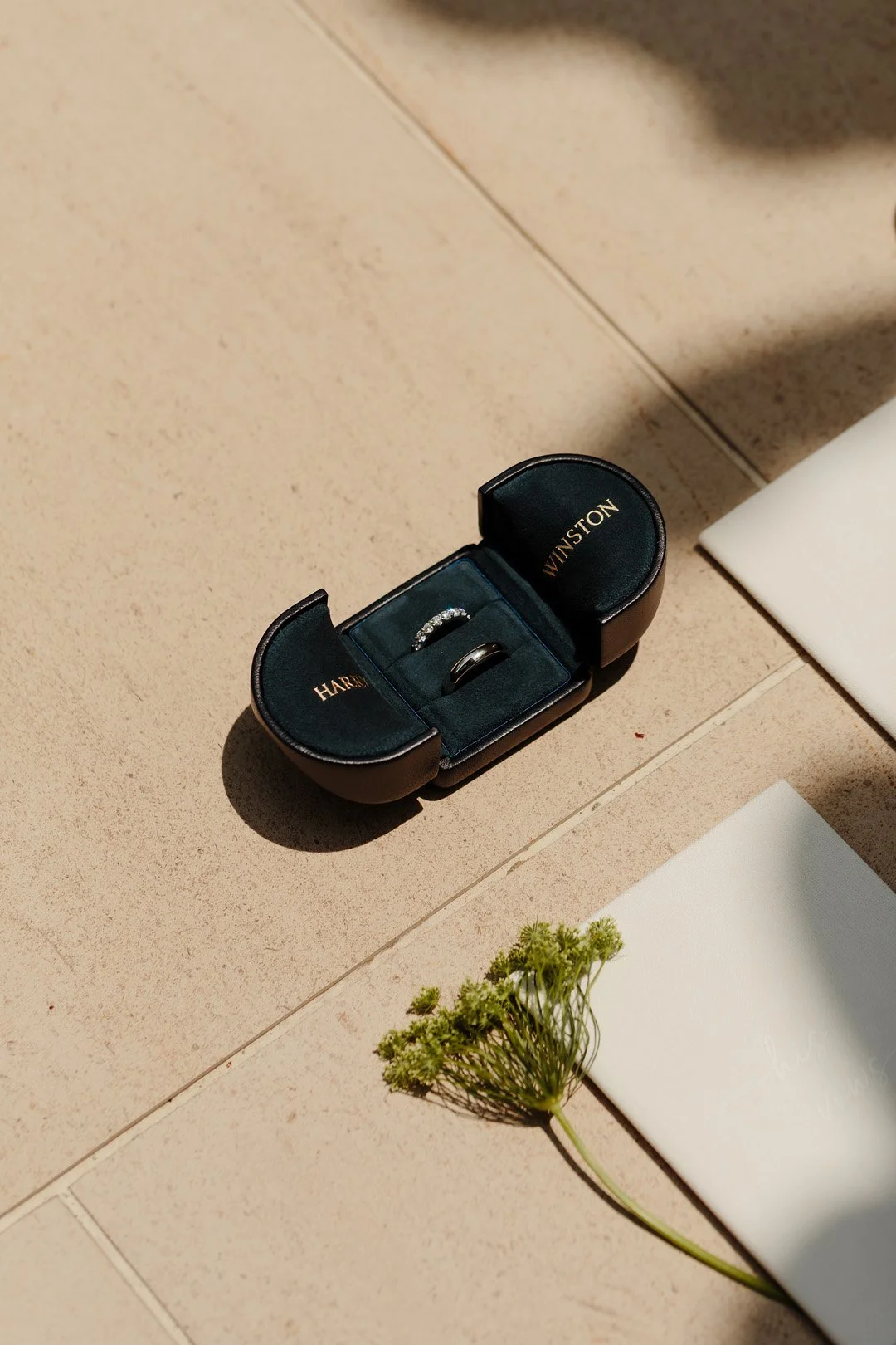An open black jewelry ring box with gold lettering on the inside of the lid, containing a diamond ring and a plain band, placed on a beige tiled surface next to a small sprig of green flowers and a white envelope.