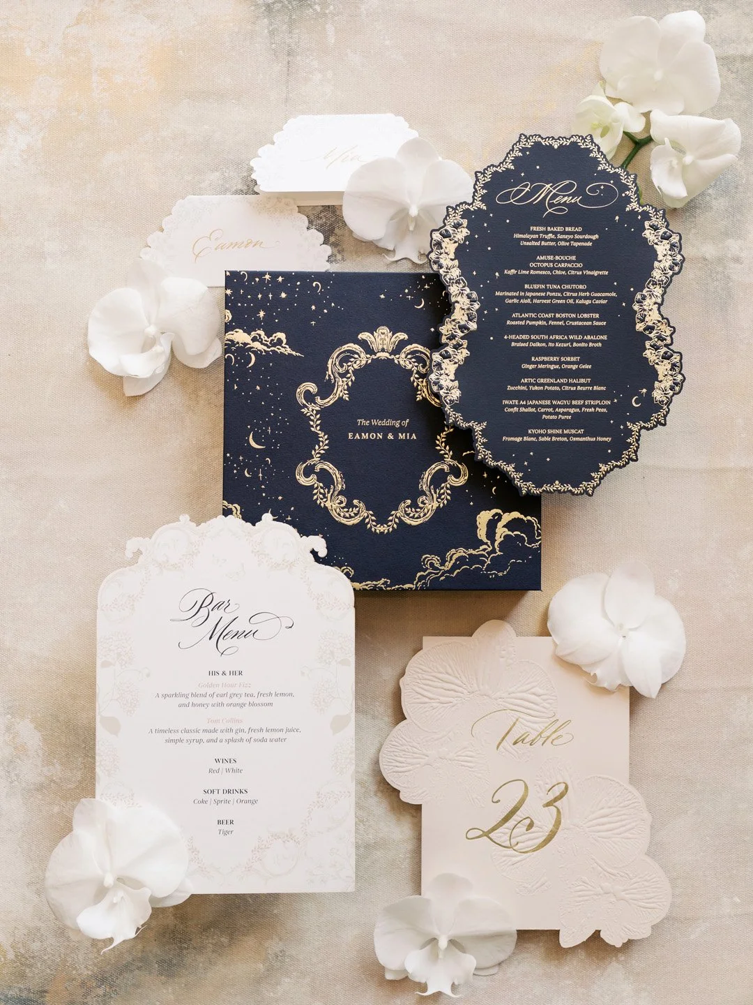 Wedding invitation suite including a main navy blue card with gold ornate border and text, a navy blue menu card with gold border and text, a white bar menu card with black text, and a table card with gold numbers, all decorated with white orchids an
