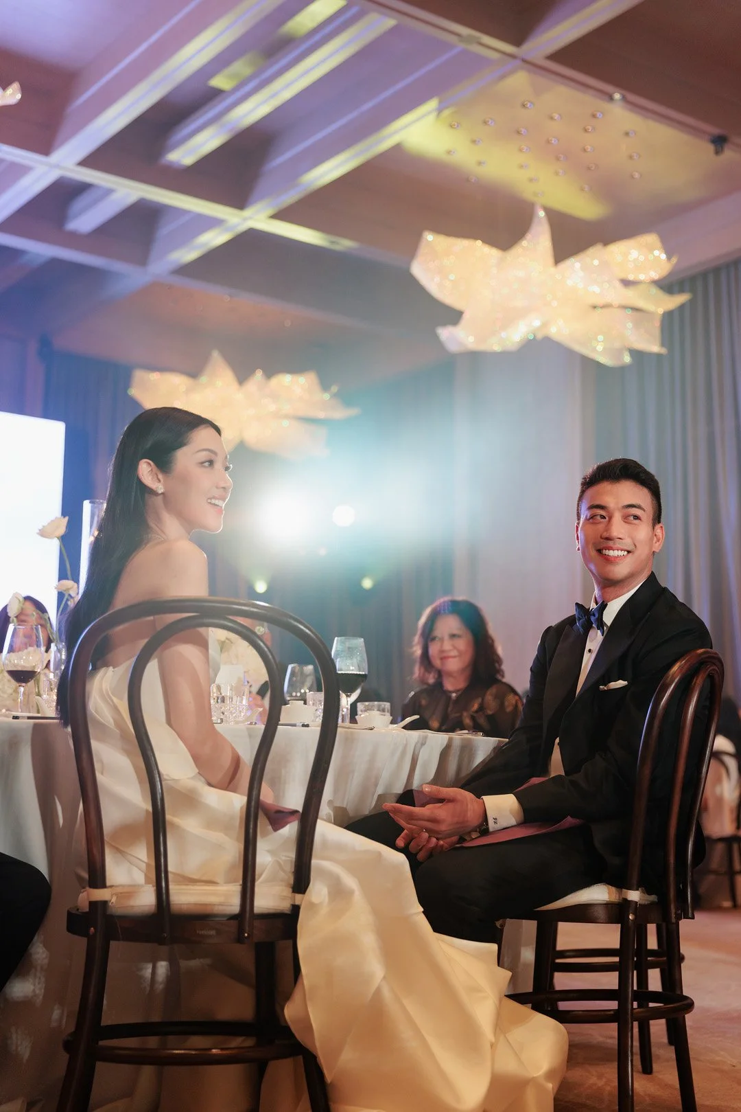 A woman in a white gown and a man in a black tuxedo with a blue bowtie sitting at a banquet table, smiling and engaging in conversation at a formal event or wedding reception. There are wine glasses and floral decorations on the table, with elegant l
