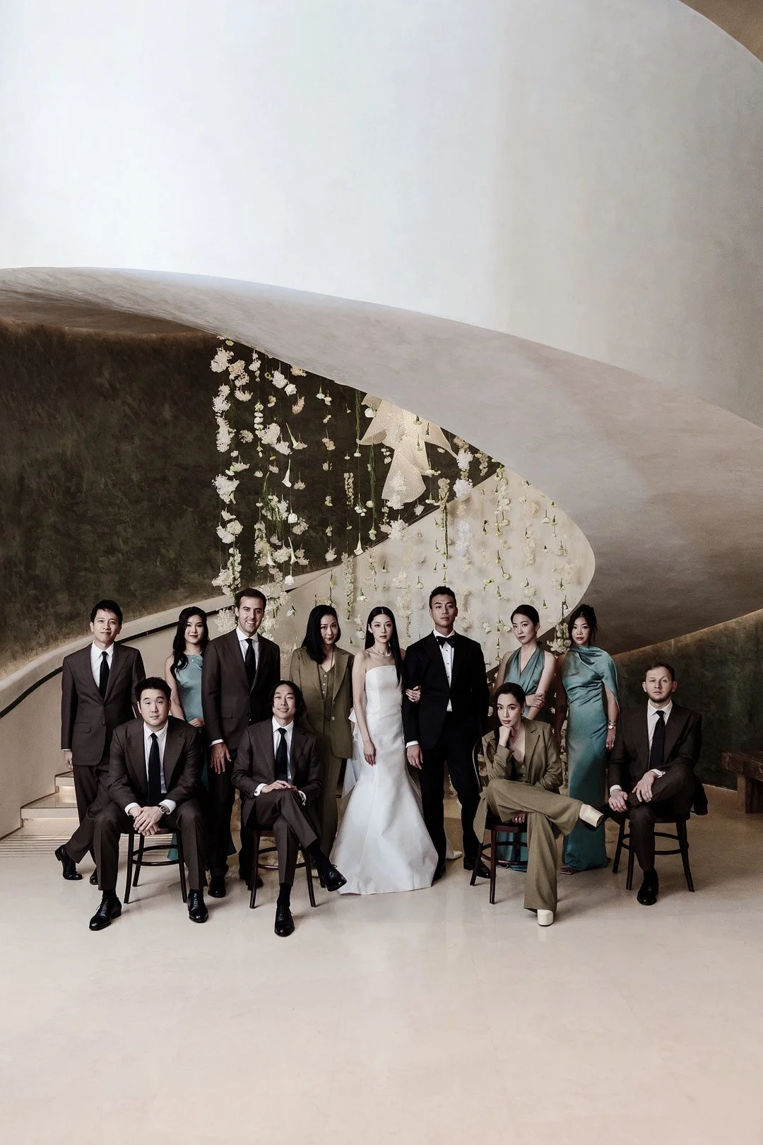 Group of people dressed in formal attire, posing in front of a modern curved wall decorated with white flowers and hanging ornaments, in an elegant indoor setting.