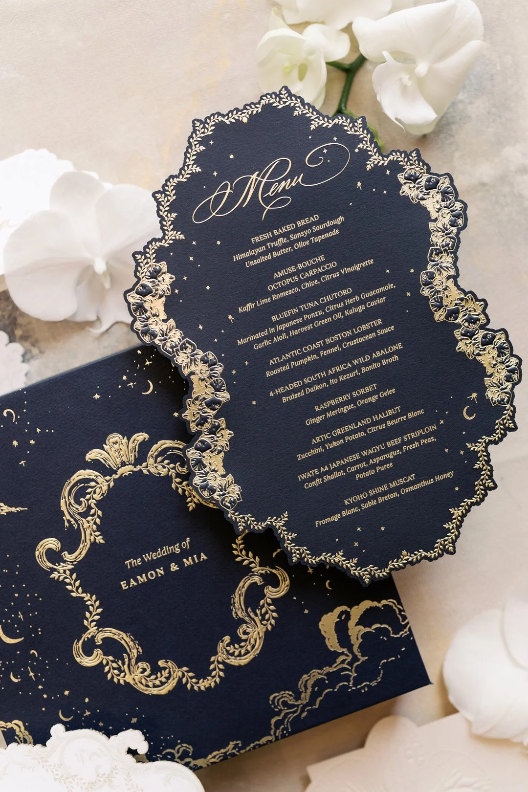 A navy blue wedding menu card with gold foil details and a matching navy blue wedding invitation with gold foil accents. The menu lists various gourmet dishes, and the invitation displays the text "The Wedding of Eamon & Mia" surrounded by decorative