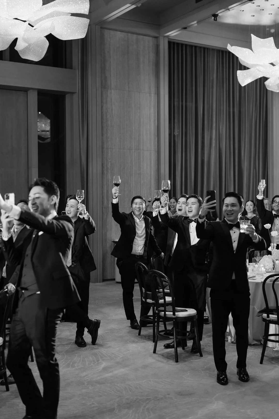 Men in tuxedos raising wine glasses in a toast at a formal event or celebration, with smiling faces in a decorated banquet room.