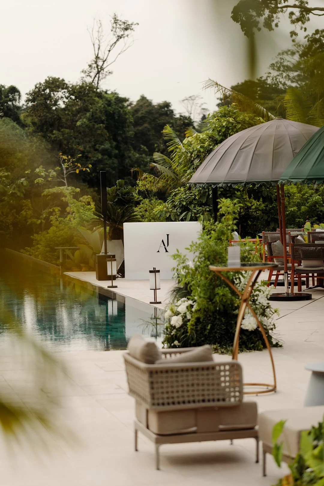 Outdoor lounge area with umbrellas, seating, and lush greenery around a pool, with a white sign displaying 'AJ' in the background.