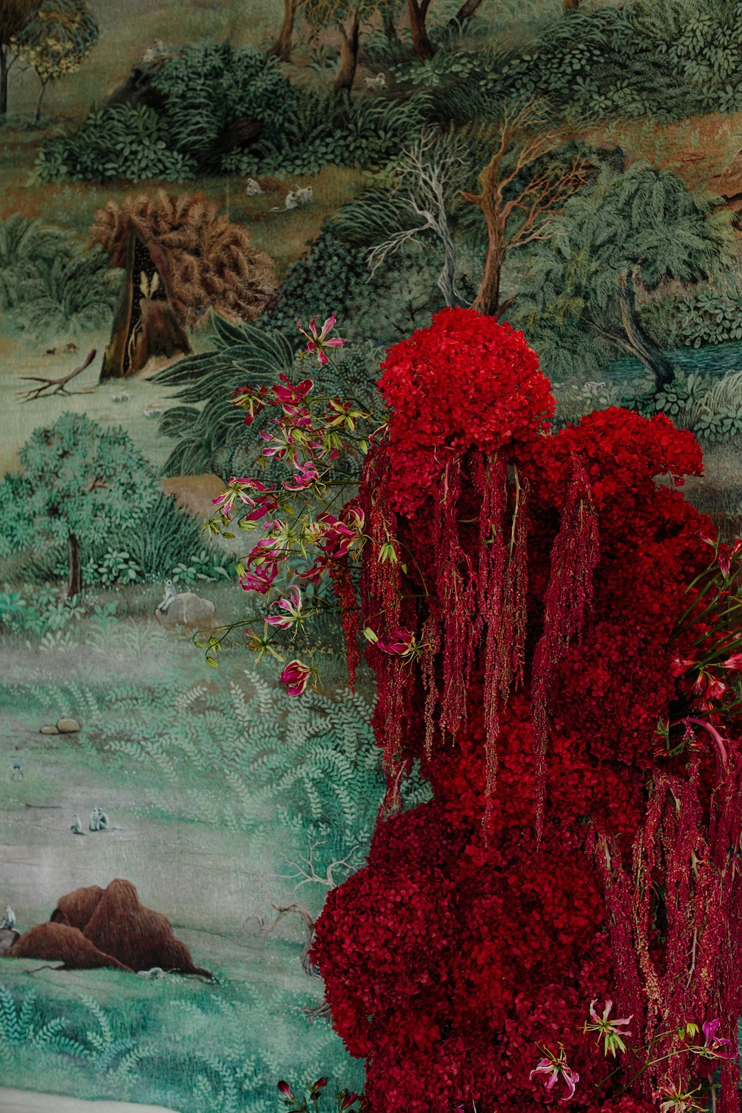 A backdrop of a lush, detailed nature scene with trees, bushes, mountains, and animals, contrasted by bright red floral arrangements in the foreground, including clusters of small red flowers and pink and purple orchids.