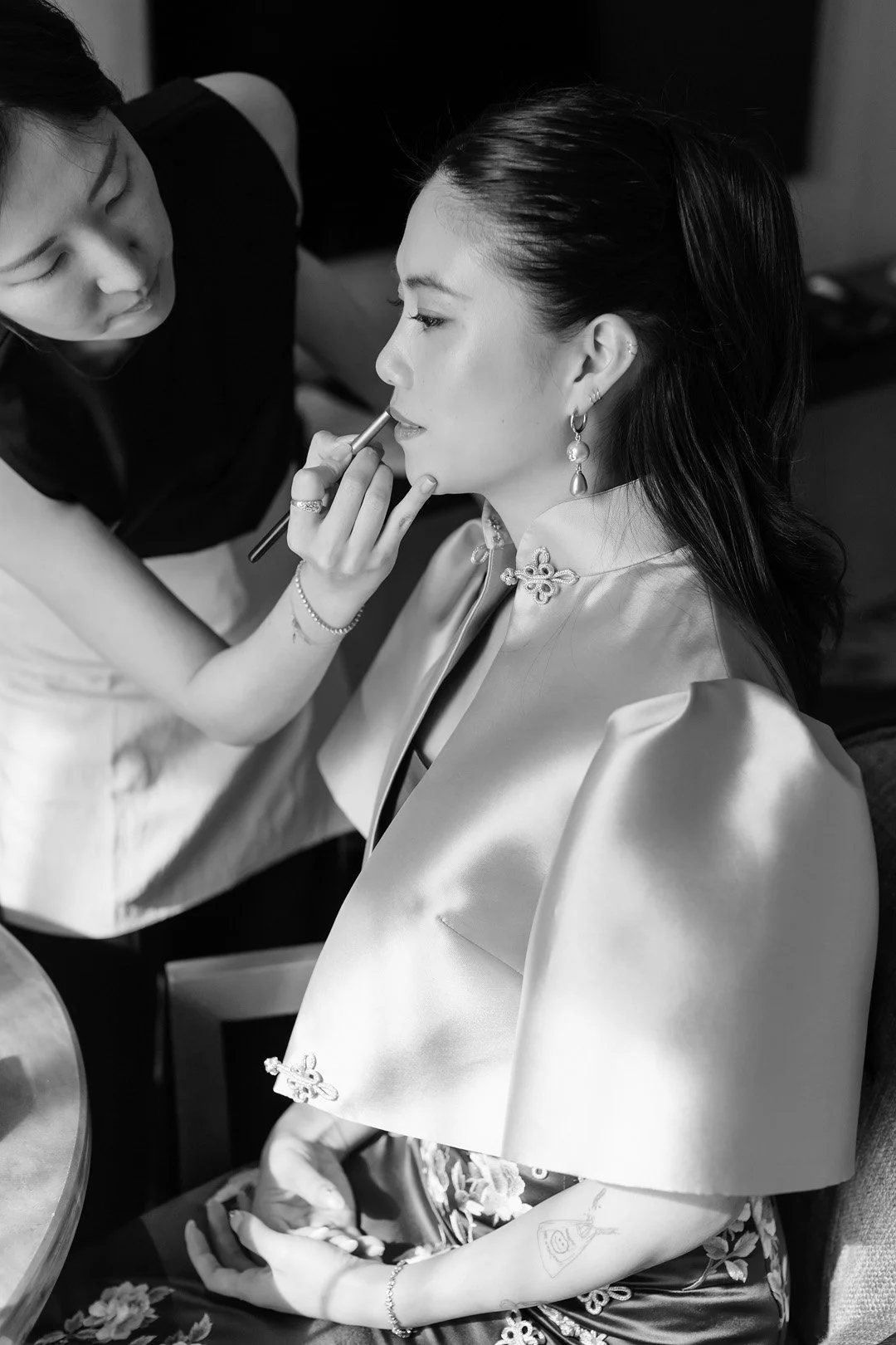 A woman with dark hair wearing jewelry and a satin jacket is getting her makeup done by a makeup artist.