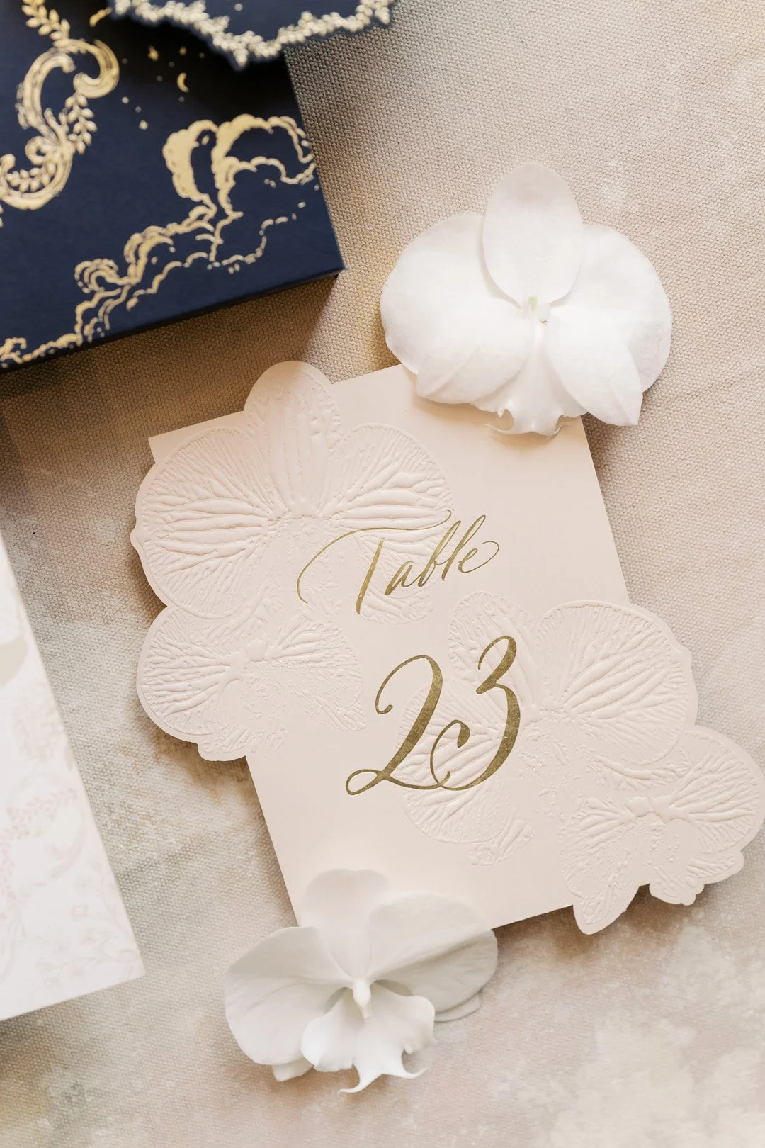 Elegant table number card with embossed butterfly design and gold script reading 'Table 23', decorated with white flower petals on a light-colored tablecloth.