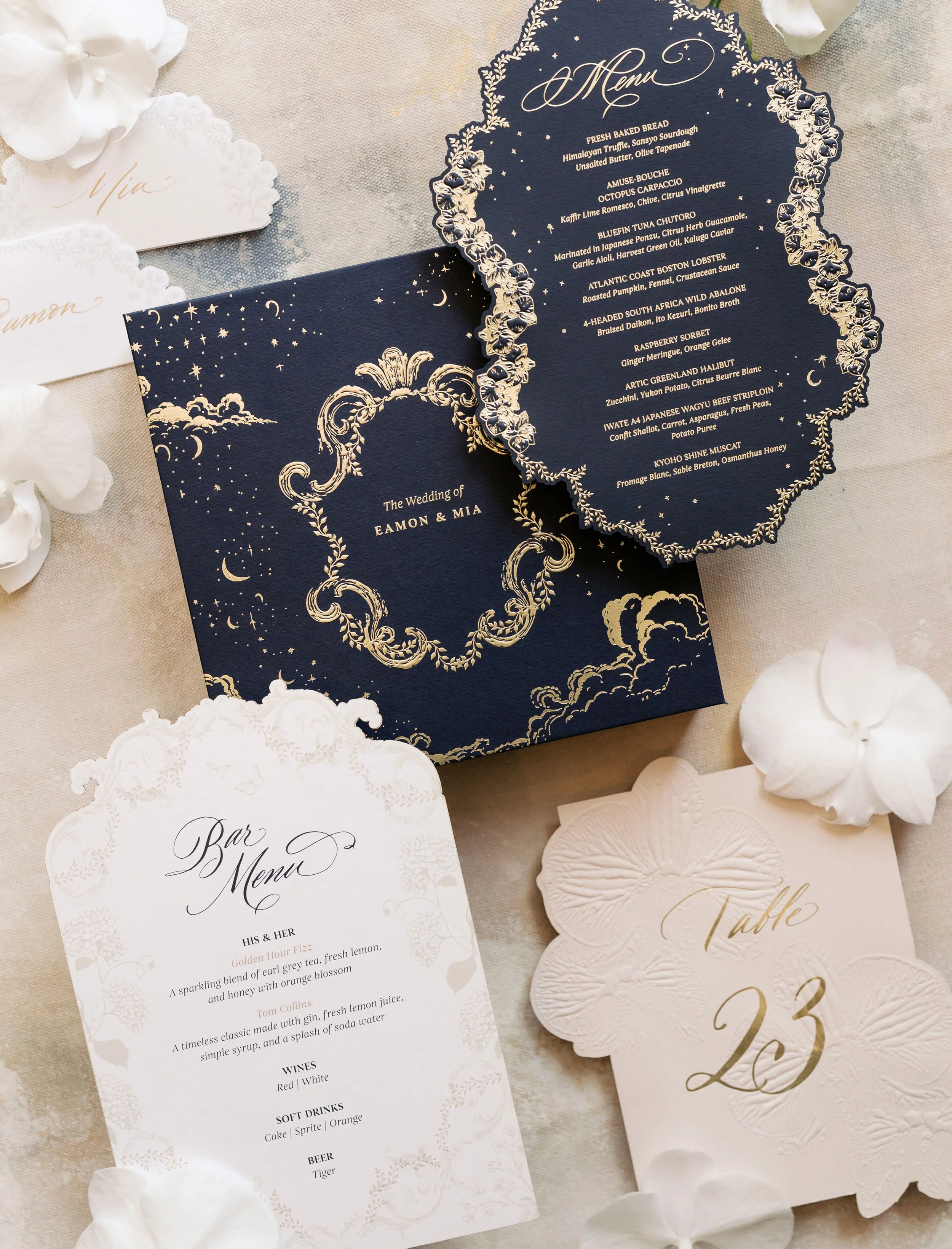 Elegant wedding stationery including a navy blue menu card with gold floral details, a wedding box with gold embroidery, a bar menu, and a table number card, all surrounded by white flowers.