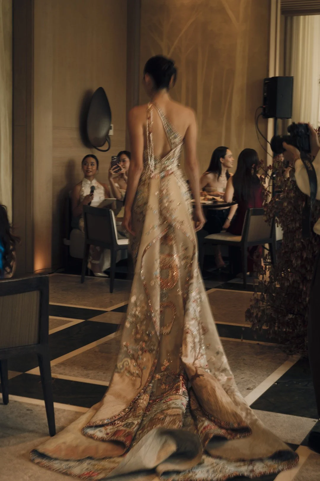 A woman in an elaborate, backless gown with intricate patterns, standing in a room with seated guests, some of whom are taking photos and holding microphones.