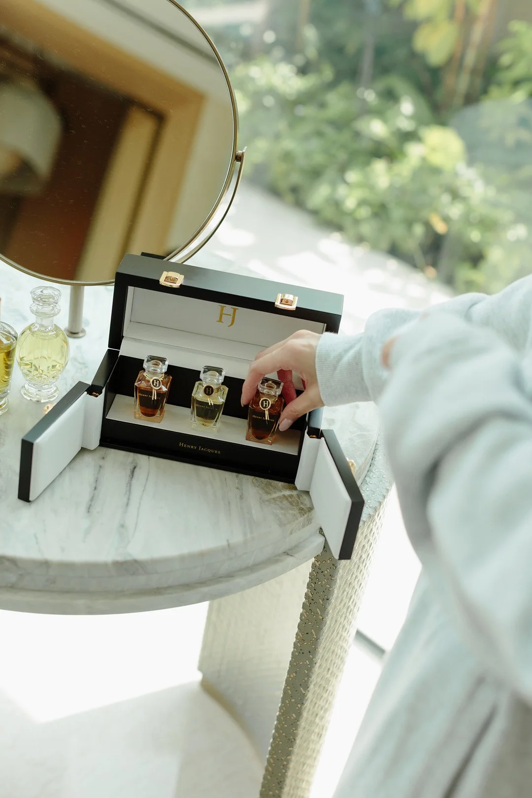 A person selecting small perfume bottles from a black and white Henry Jacques perfume box on a marble table, with a round mirror and other perfume bottles nearby.