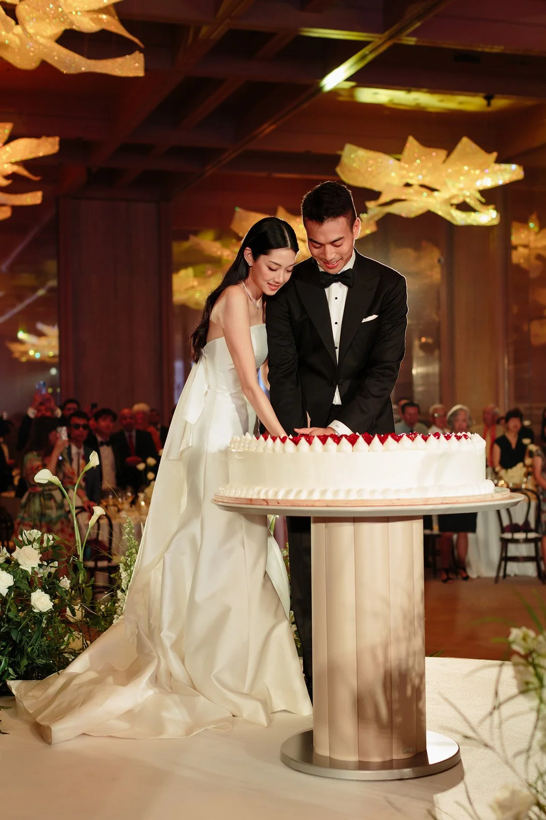 A bride and groom cutting a wedding cake together at their wedding reception, surrounded by seated guests and elegant floral arrangements.