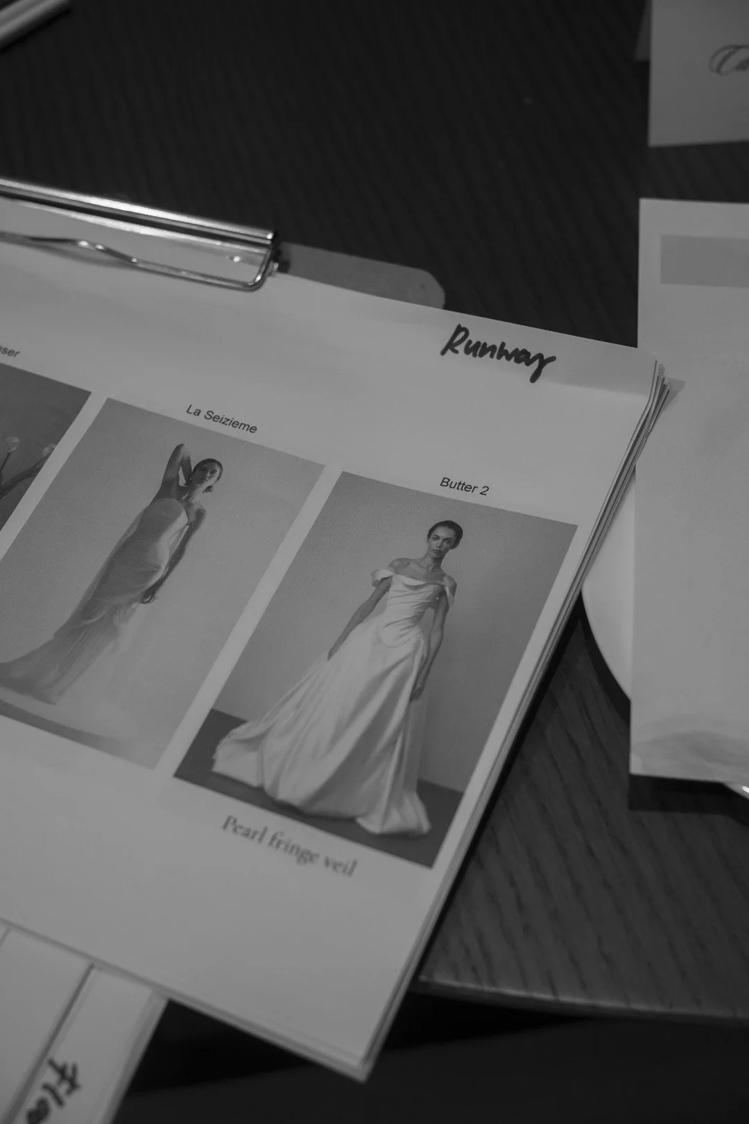 A black and white photo of a fashion design sketchbook with images of wedding dresses, labeled 'La Seizieme', 'Butter 2', and 'Pearl Fringe velvet'. The word 'Runway' is handwritten at the top. The sketchbook is held in place by a metal clip, resting